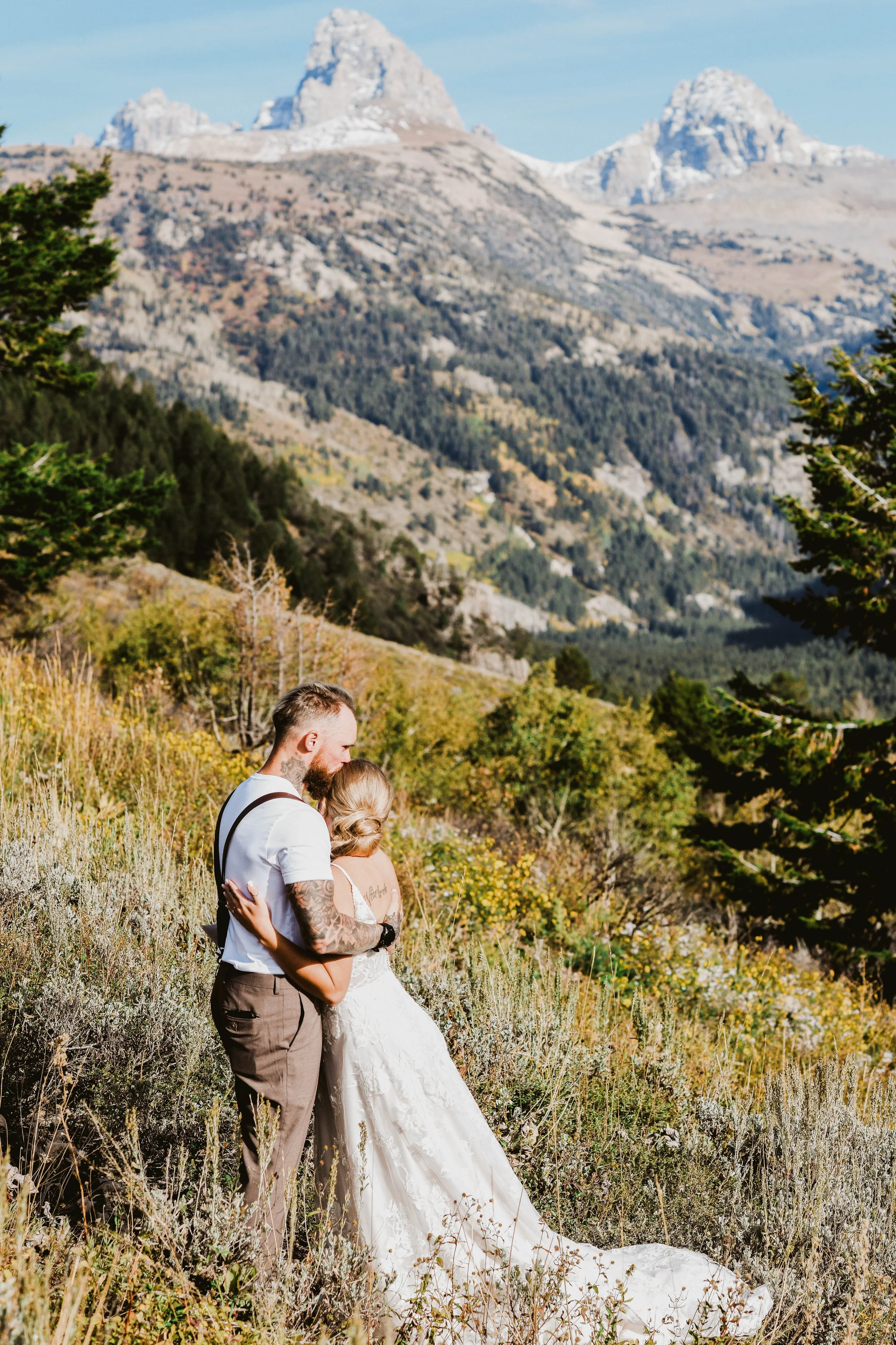 A couple on their wedding day embracing in a scenic mountain landscape with snow-capped peaks and green forest in the background.