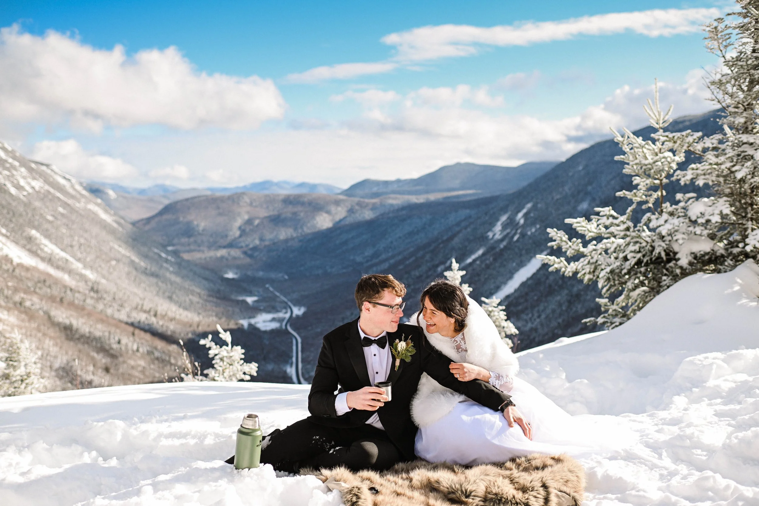 A newlywed couple in wedding attire sitting on a blanket in the snow on a mountain with a scenic view of snowy mountains and a blue sky in the background.