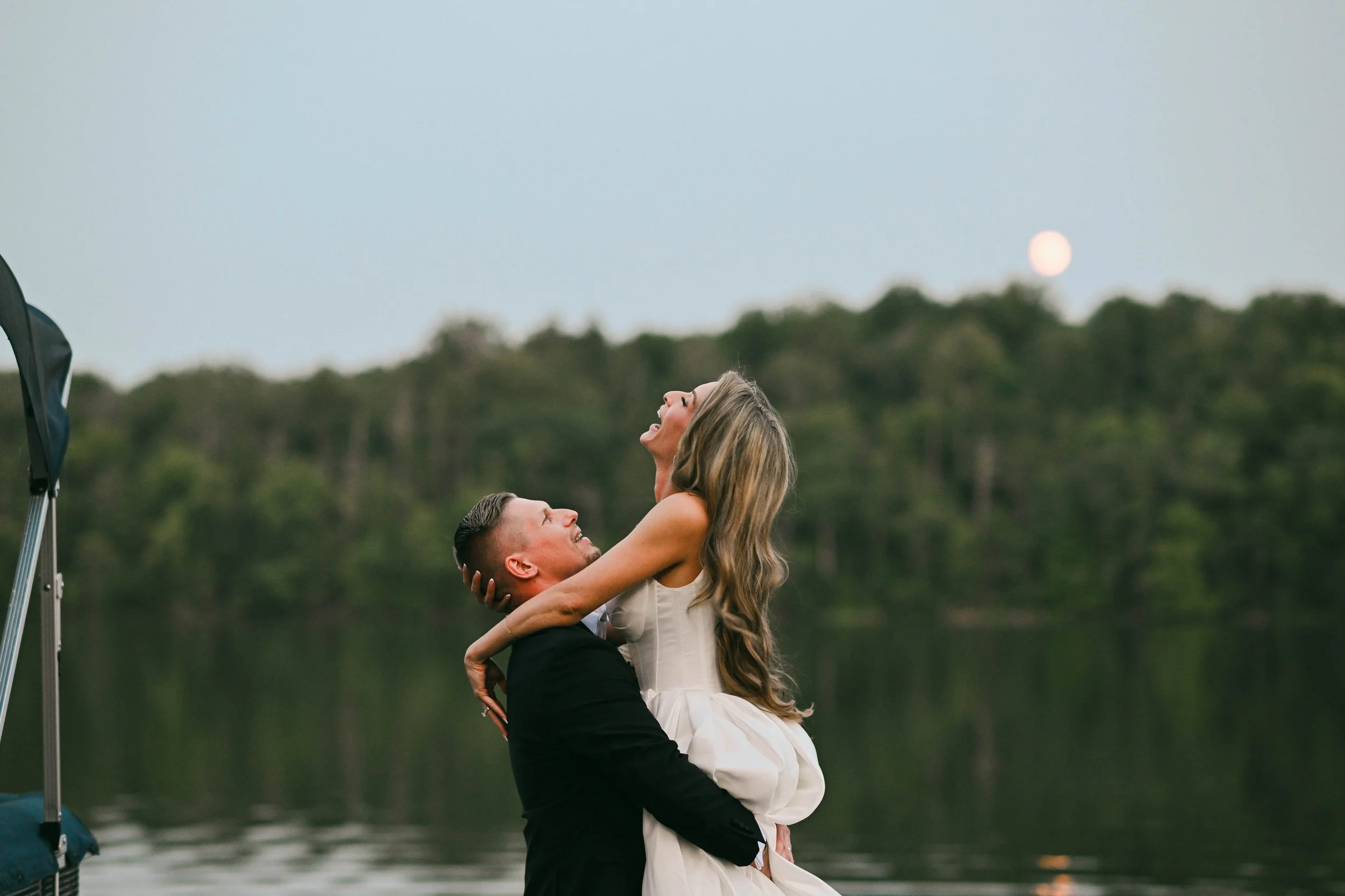 A man in a black suit lifting a woman in a white dress by a lakeside at dusk, with the moon rising in the background, both smiling and enjoying the moment.