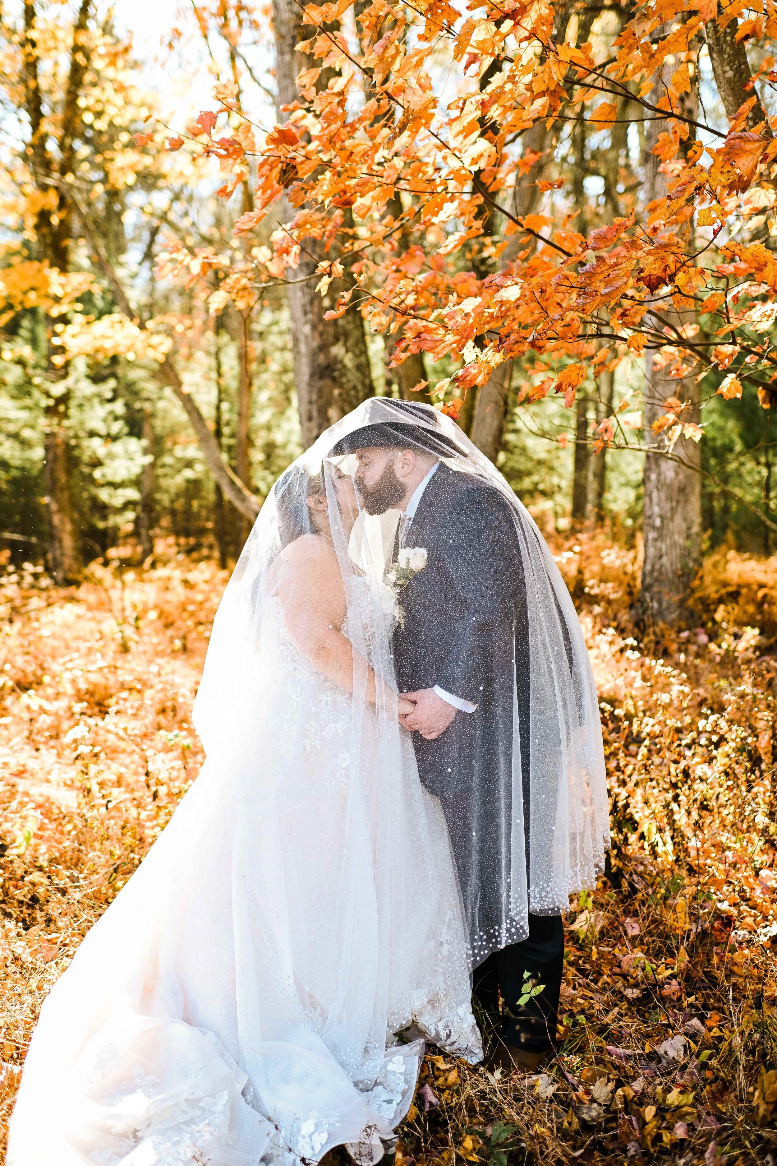 A bride and groom share a kiss under a veil in a forest with autumn leaves.