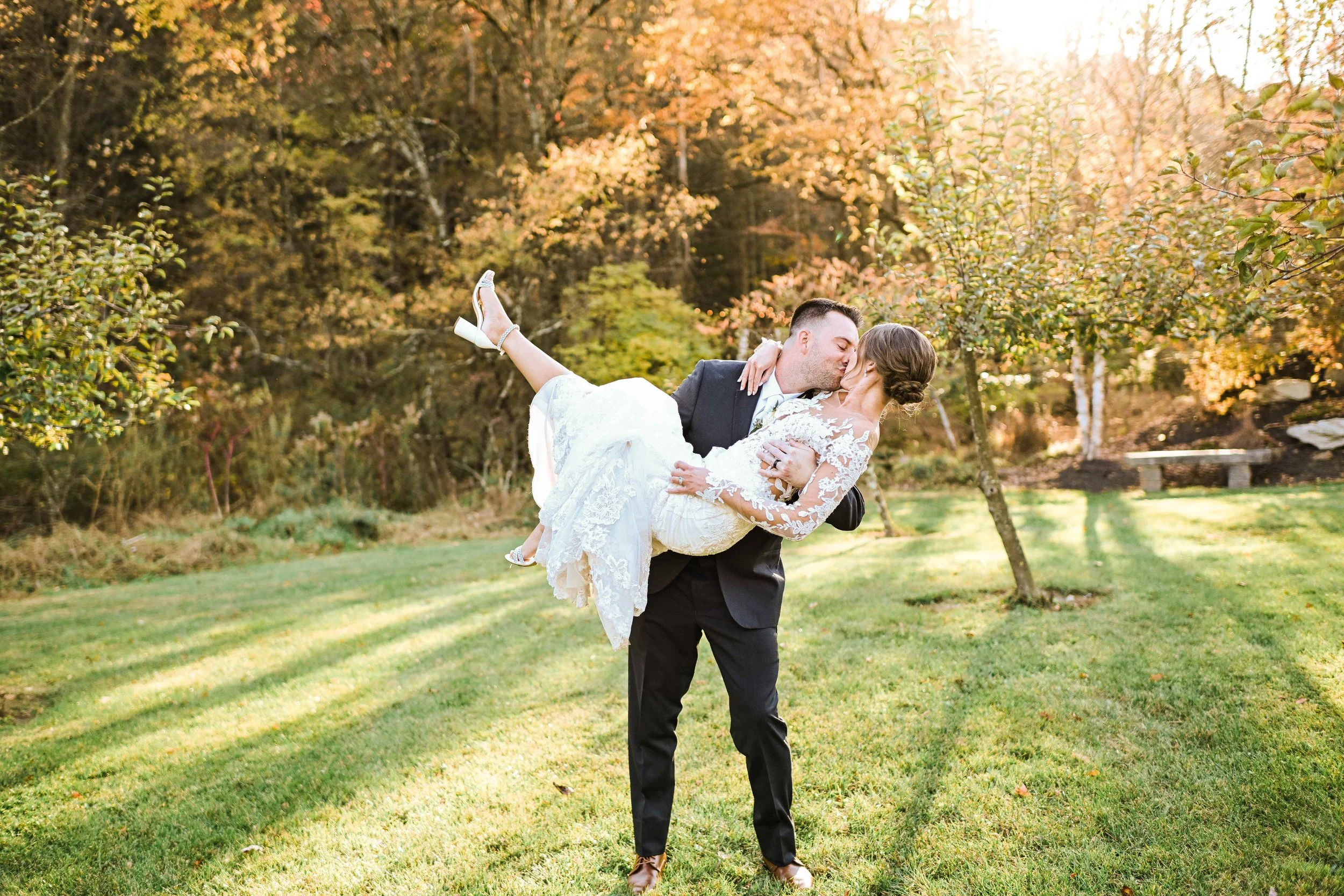 A groom in a black suit lifts and kisses a bride in a white lace dress outdoors on a grassy field during autumn, with trees and sunlight in the background.