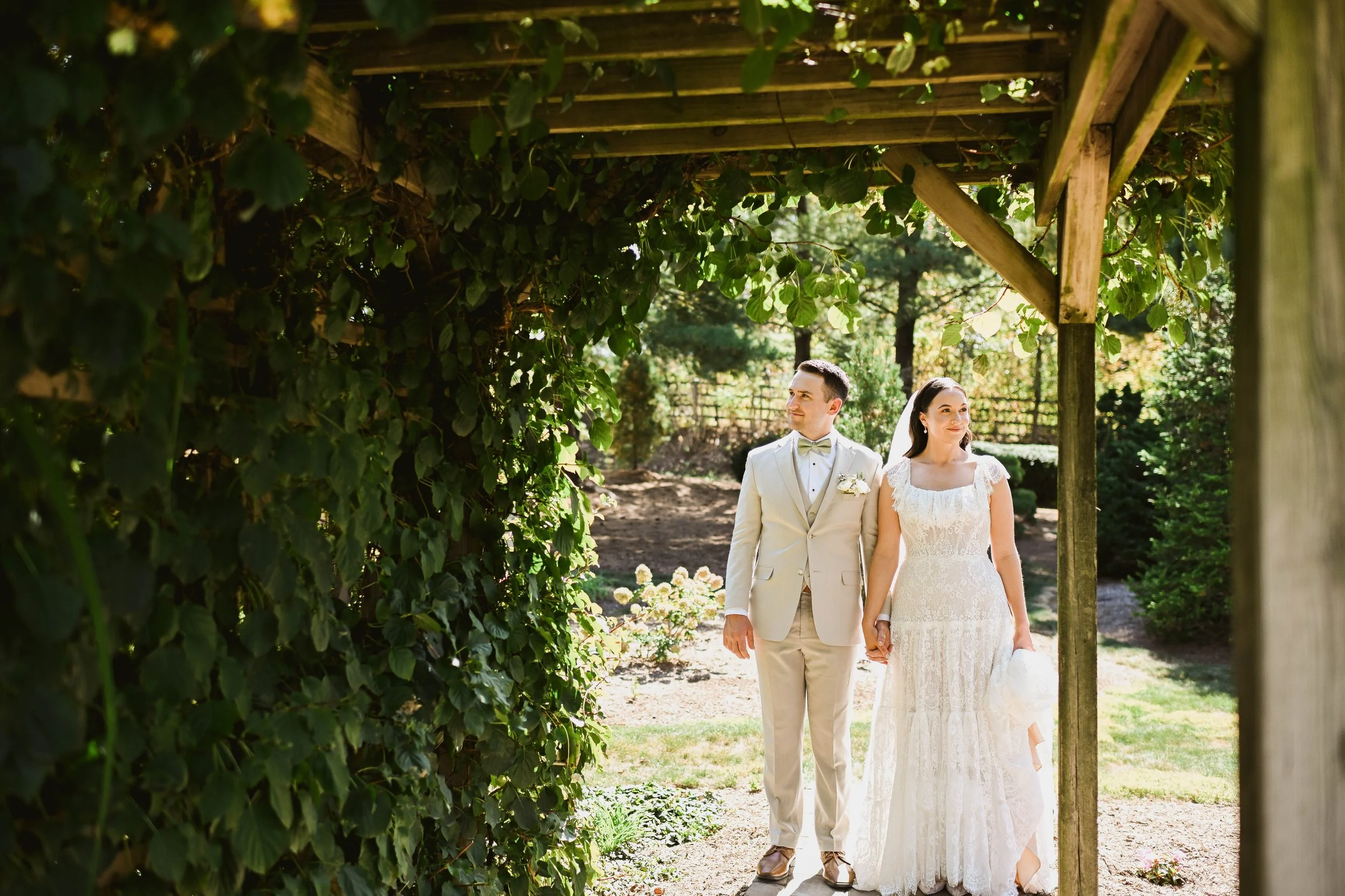 A bride and groom holding hands under a wooden pergola with greenery, in an outdoor wedding setting on a sunny day.