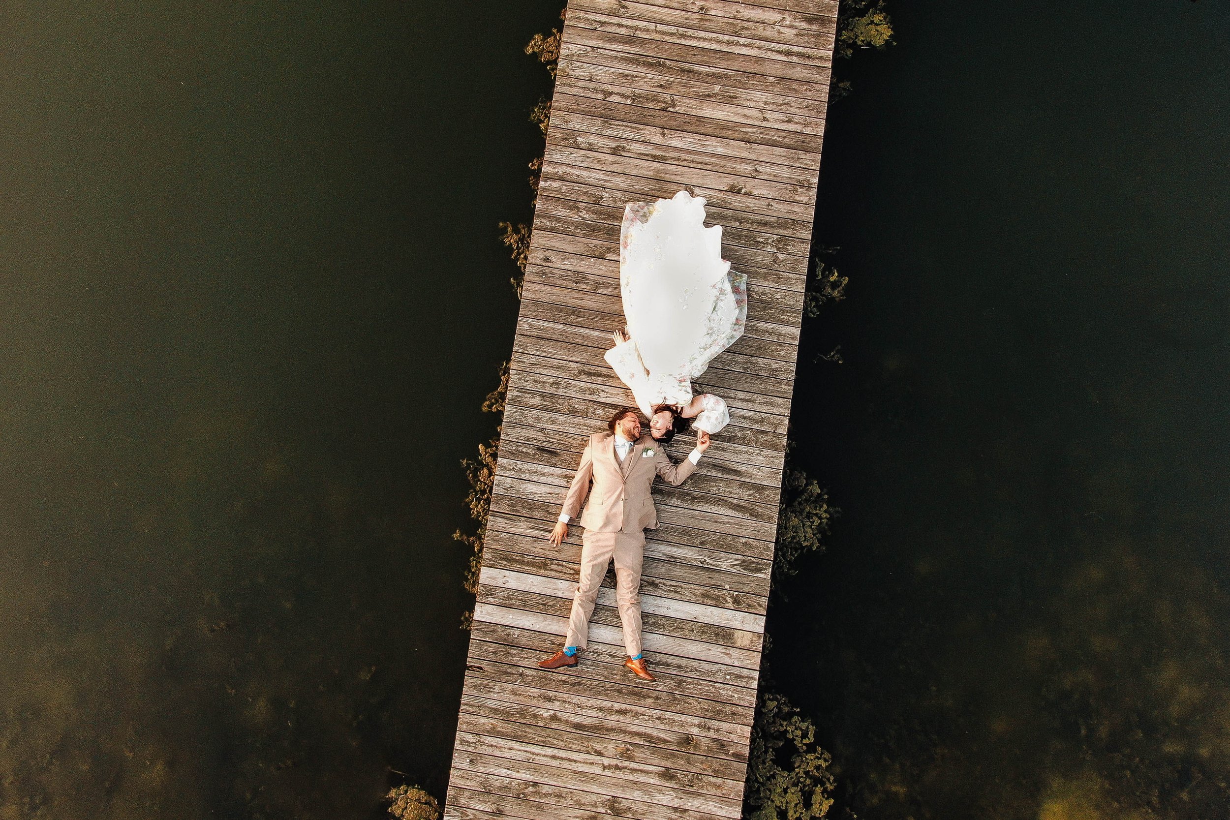 A bride and groom lying on a wooden dock over water, holding hands and facing each other, with a wedding dress spread out beside them.