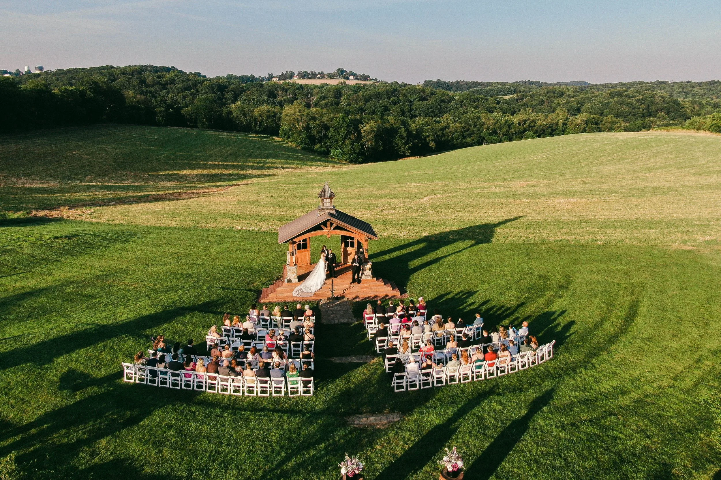 An outdoor wedding ceremony taking place on a grassy field with a small wooden chapel, surrounded by white chairs filled with guests, and a bride and groom standing under the chapel's roof with the officiant, all set against a backdrop of rolling hil