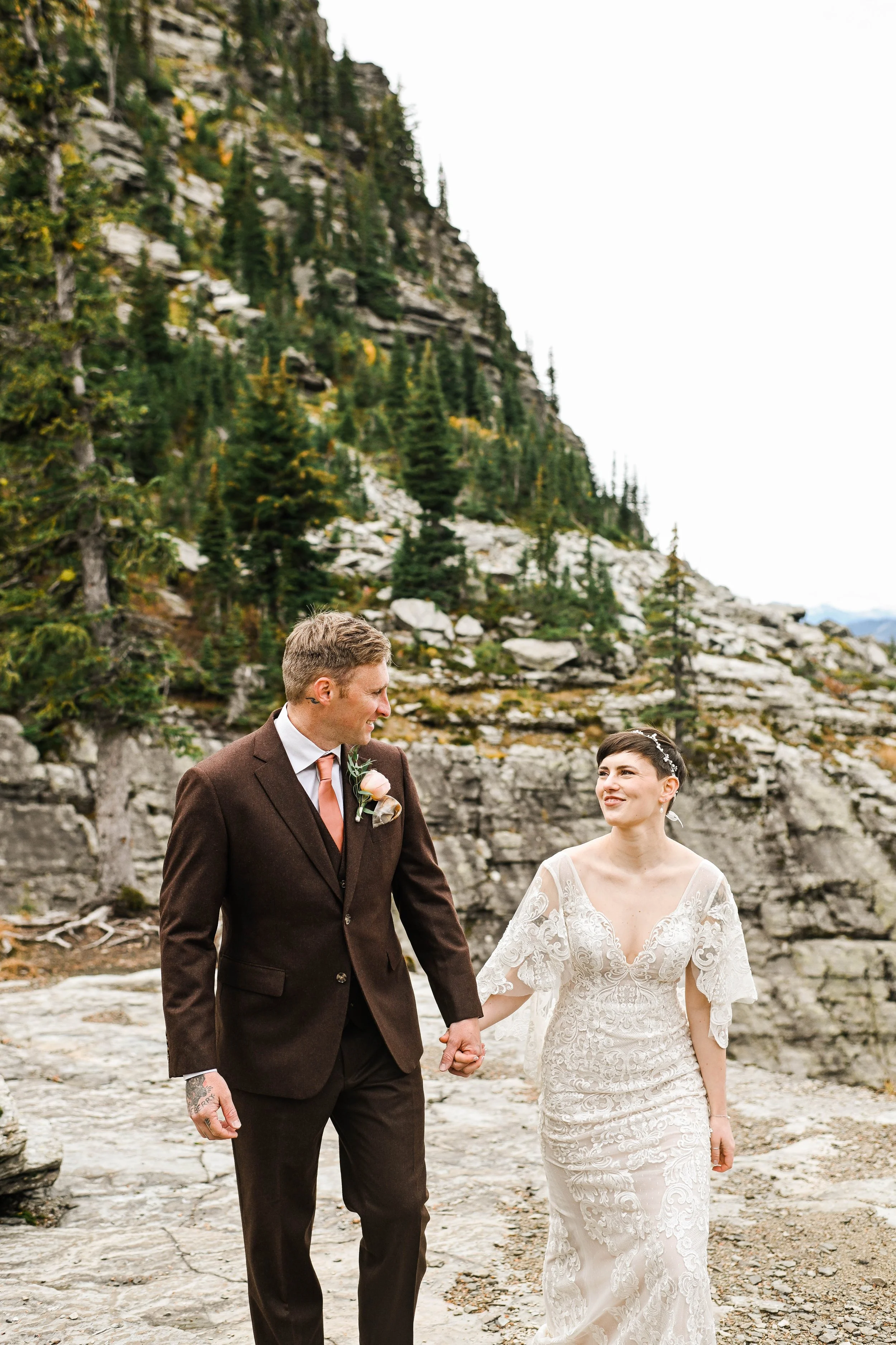 A bride and groom holding hands and walking outdoors near rocky terrain and evergreen trees, with a mountain in the background.
