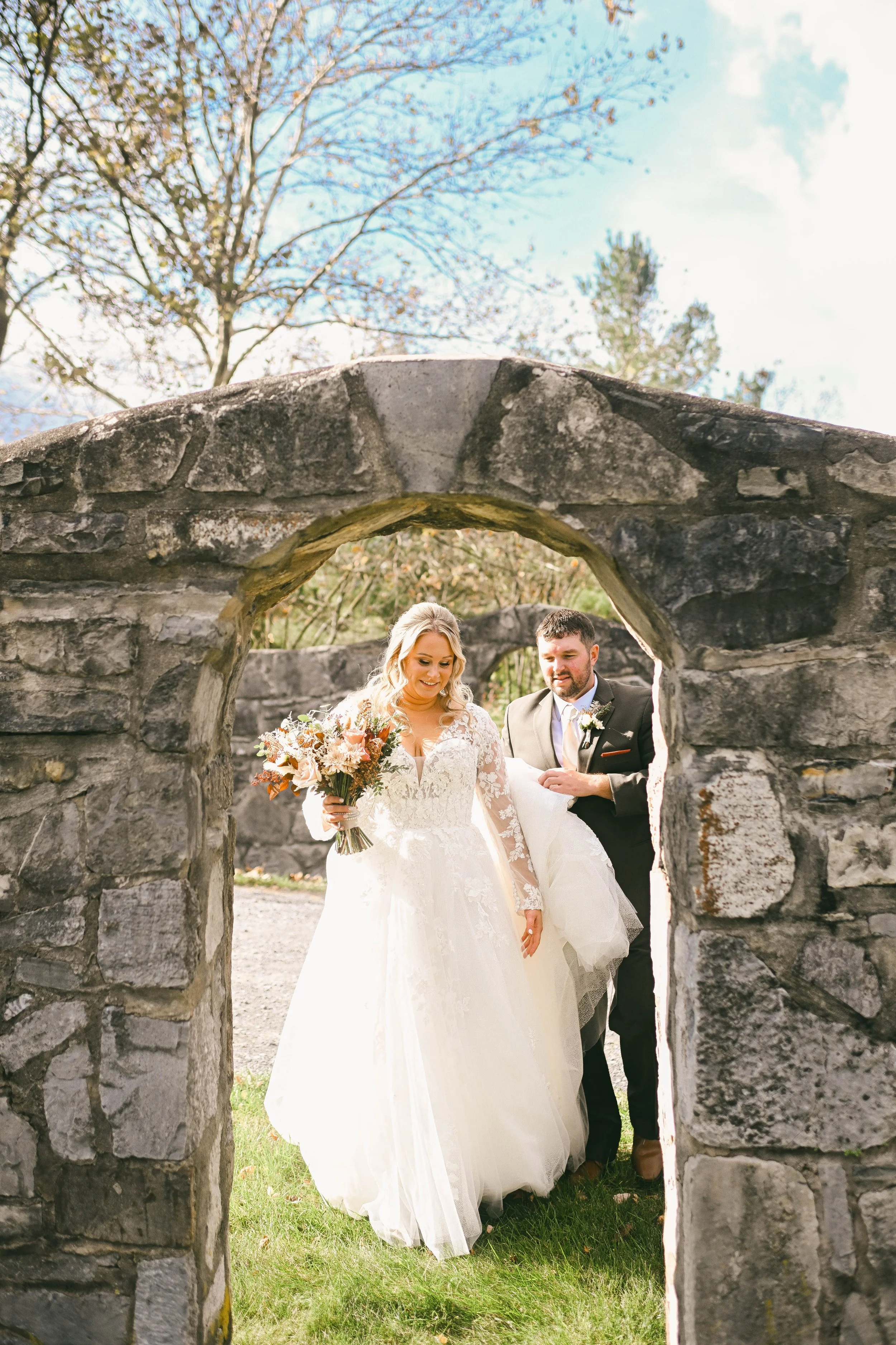 Bride in a white wedding dress holding a bouquet of flowers, walking through a stone archway, with a groom in a dark suit behind her, outdoors on a sunny day.