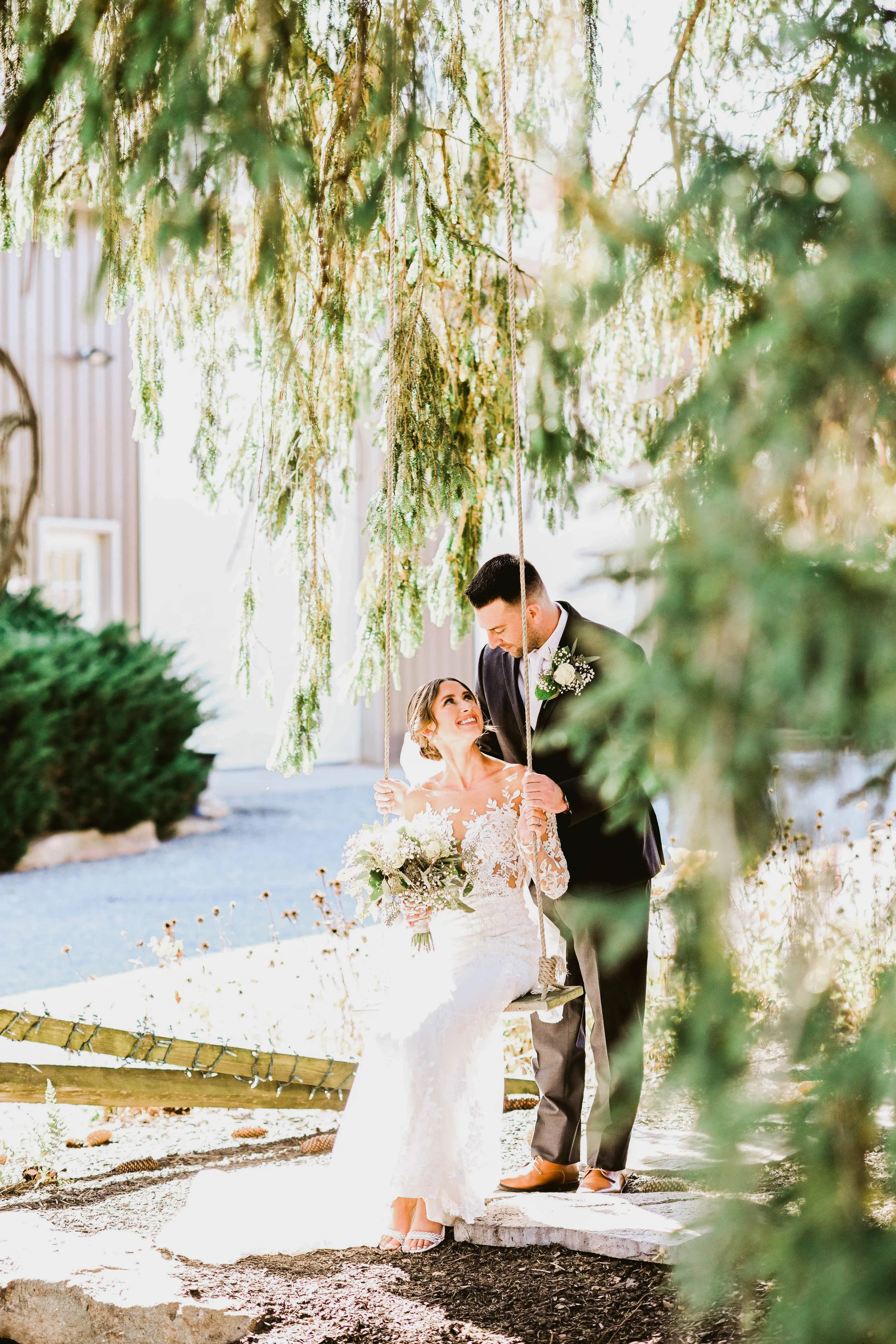 A bride and groom on their wedding day, standing on a small platform under a swing made of vines and hanging greenery. The bride is holding a bouquet and smiling at the groom, who is looking down at her affectionately. The scene is outdoors in natura