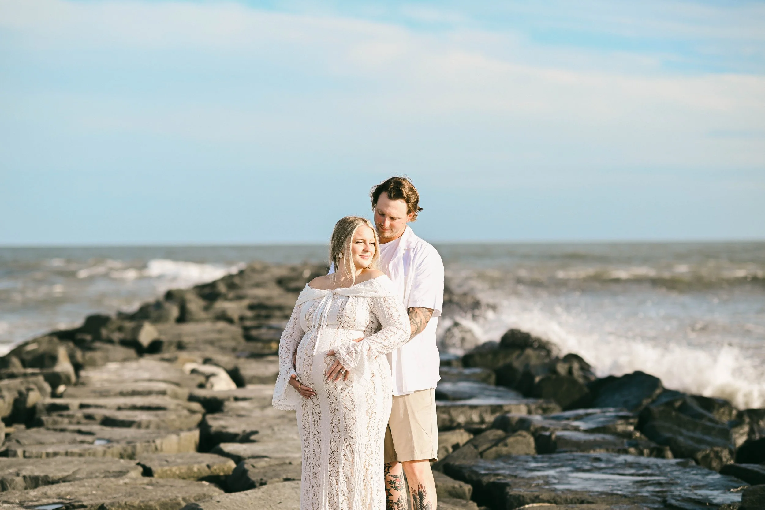 A pregnant woman and a man are standing on a rocky shoreline with the ocean and blue sky in the background. The woman is wearing a white lace dress, and the man is in a white shirt and beige shorts, holding her and looking at her affectionately.