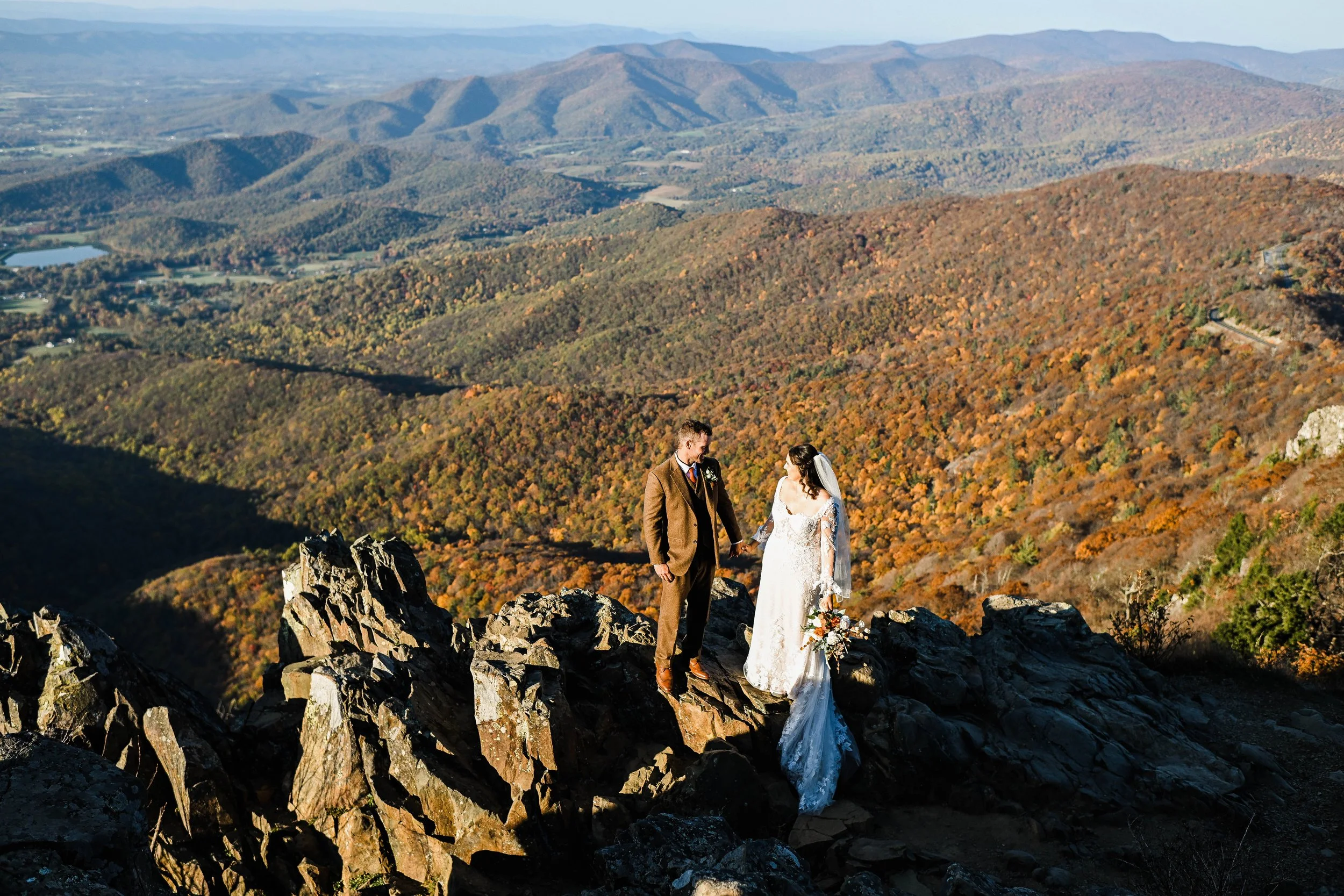 A bride and groom holding hands on a rocky mountaintop with a panoramic view of colorful autumn trees and distant mountains.
