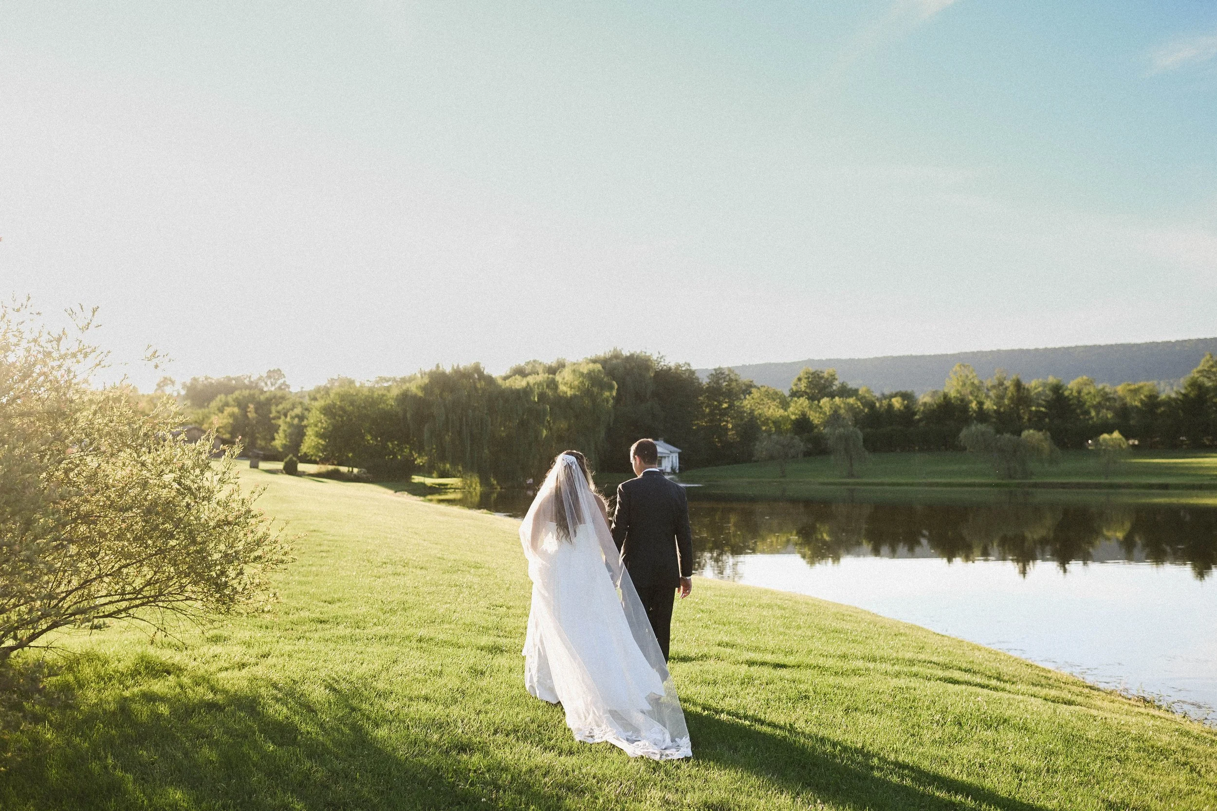 A bride and groom walking by a lake on a sunny day with trees and hills in the background.