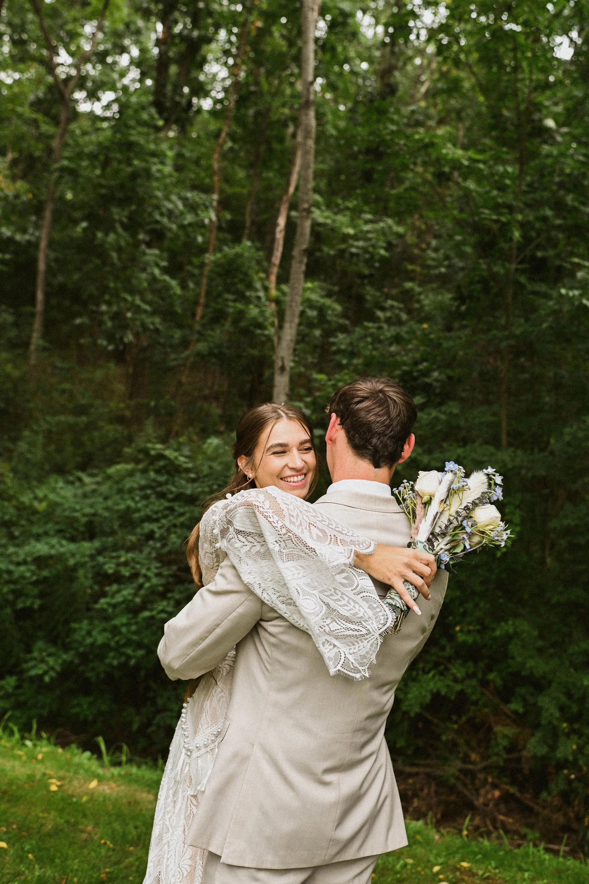 A couple is happily embracing outdoors surrounded by green trees and foliage. The woman, with long brown hair, is smiling while holding a bouquet, as the man, with short dark hair, faces her in an affectionate pose.