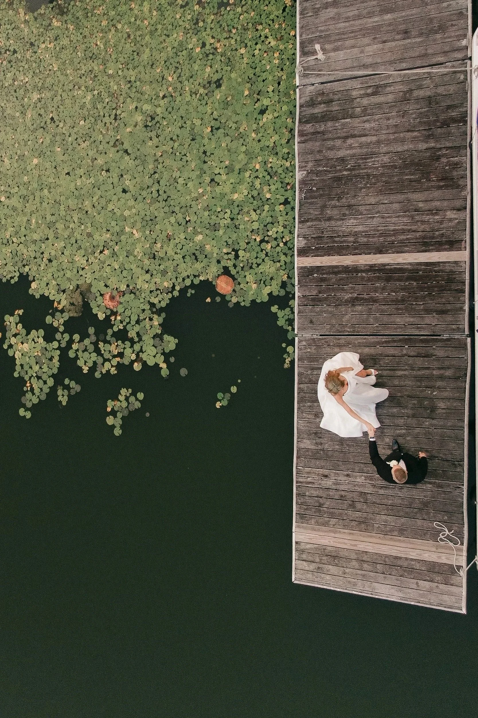 A bride and groom dancing on a wooden dock by a pond with lily pads. The bride wears a white dress, and the groom wears a dark suit. They are holding hands and dancing.
