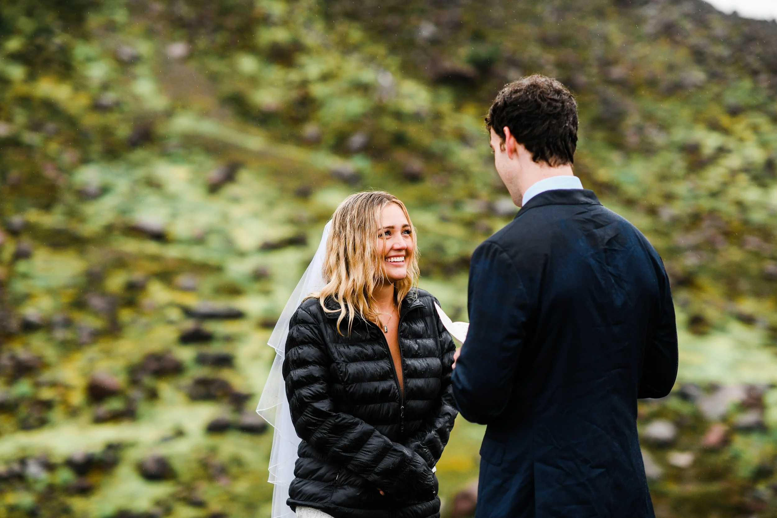 A woman at an outdoor wedding ceremony, smiling as she exchanges vows with a man who is facing her, in a natural green setting.