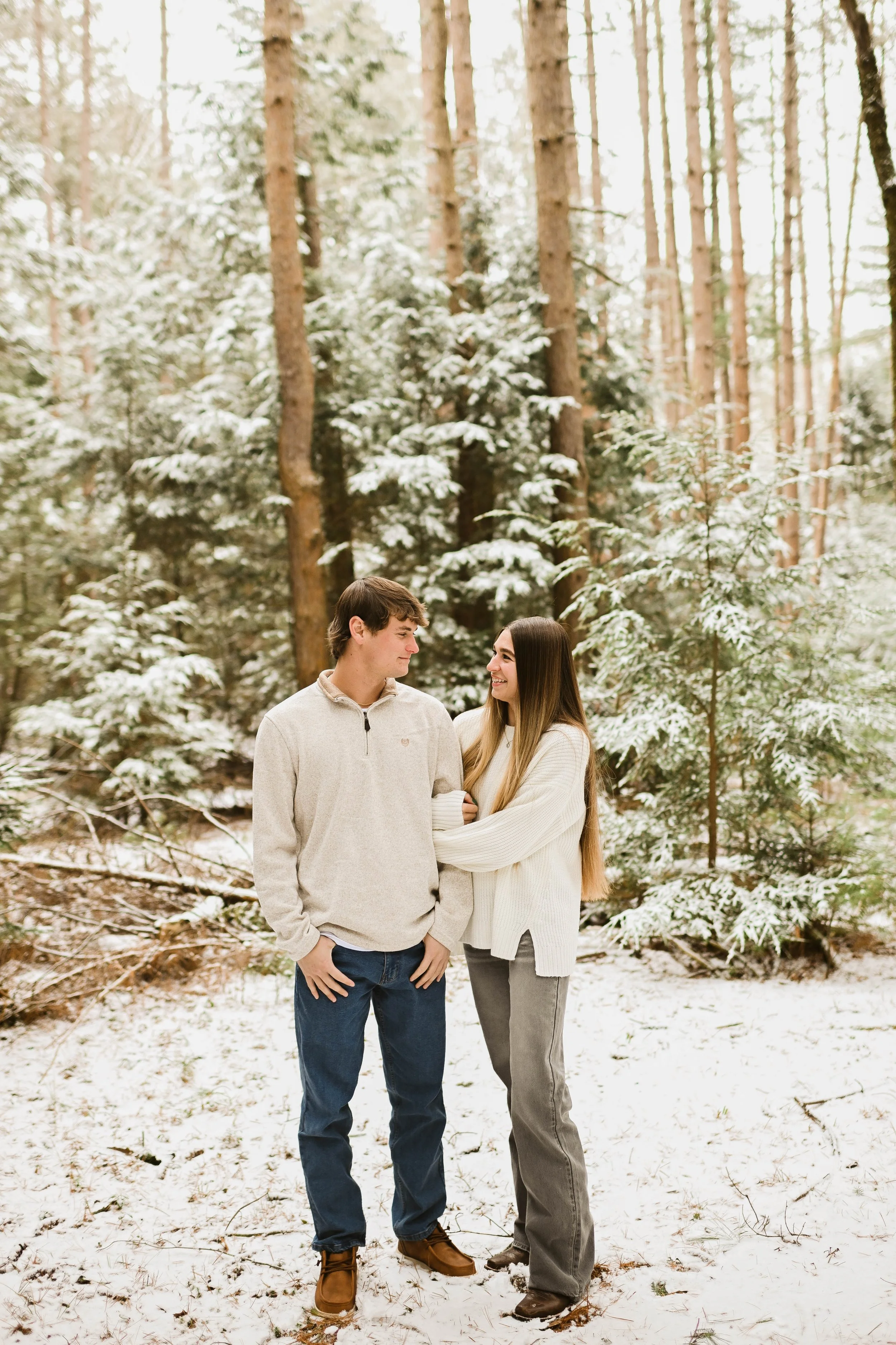 A young couple stands in a snow-covered forest, facing each other and smiling.
