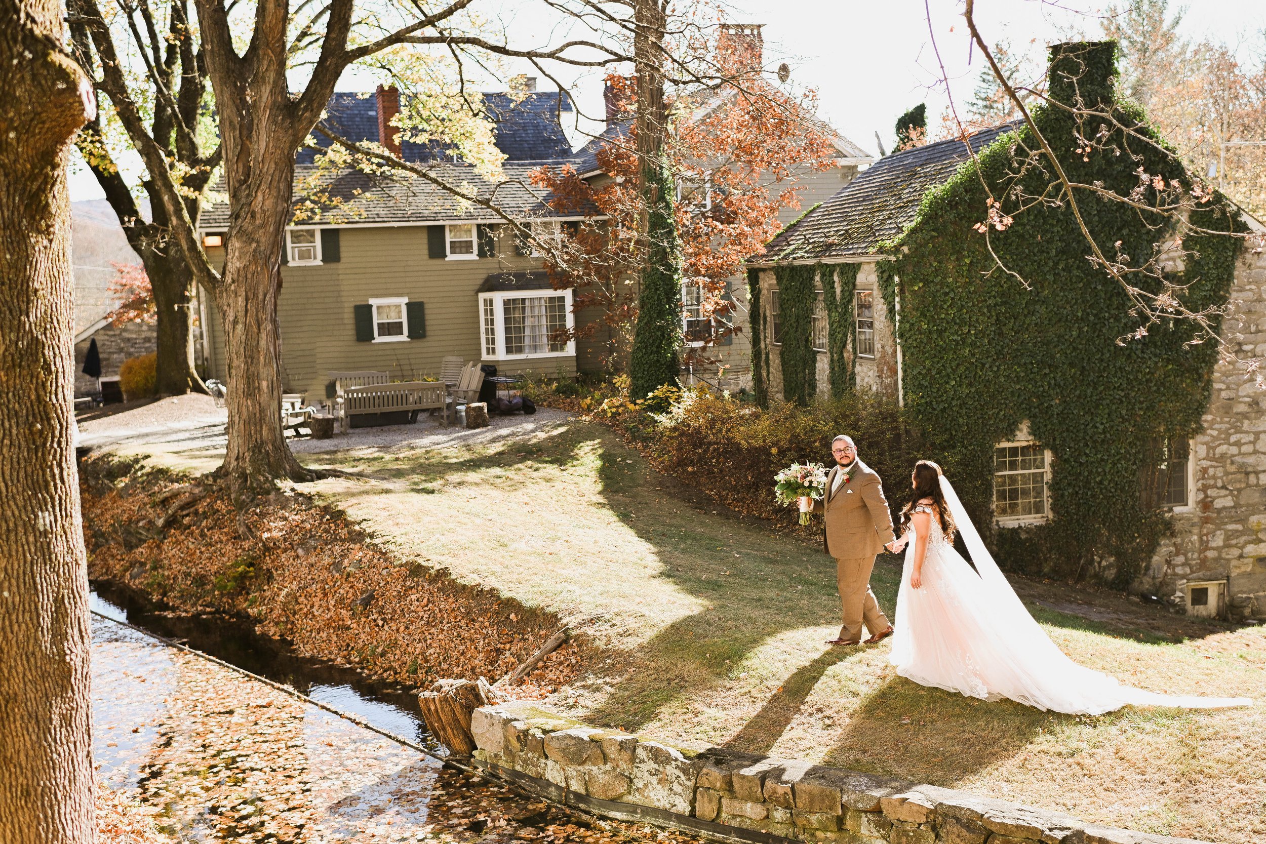 A newlywed couple walking hand in hand along a grassy pathway next to a small stream in a garden, with the bride in a white wedding gown and the groom in a brown suit holding a bouquet of flowers.