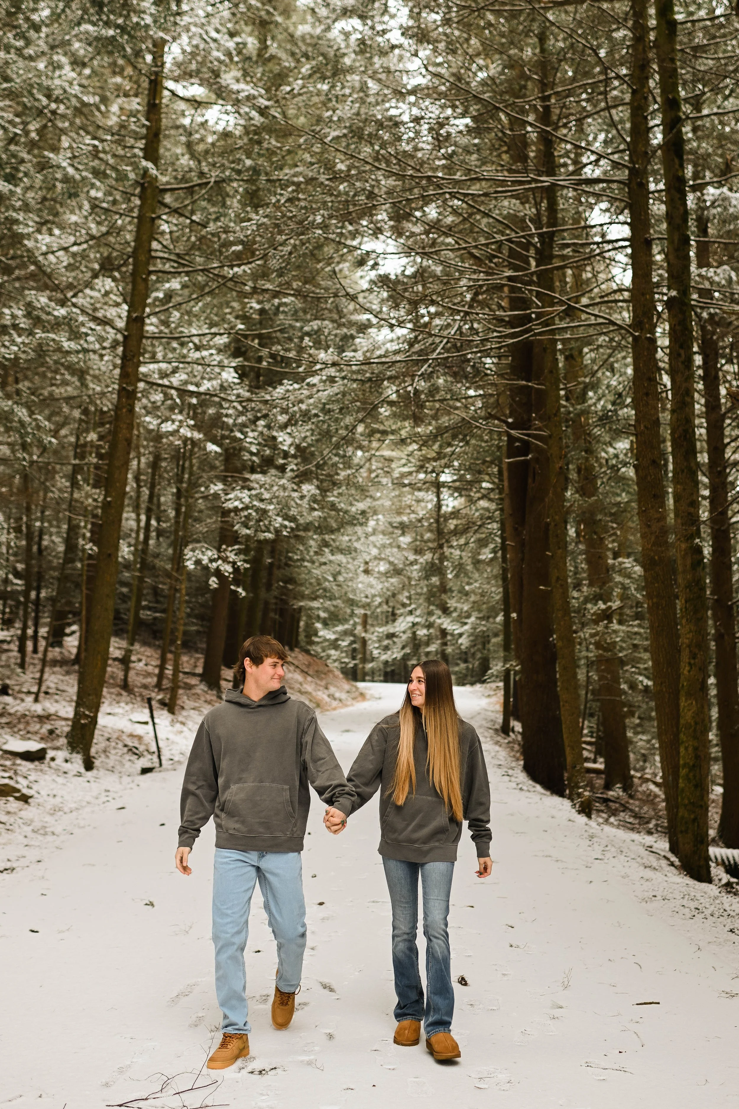A young couple walks hand-in-hand through a snow-covered forest trail during winter. They are both wearing gray hoodies and jeans, smiling at each other amidst tall, snow-dusted trees.
