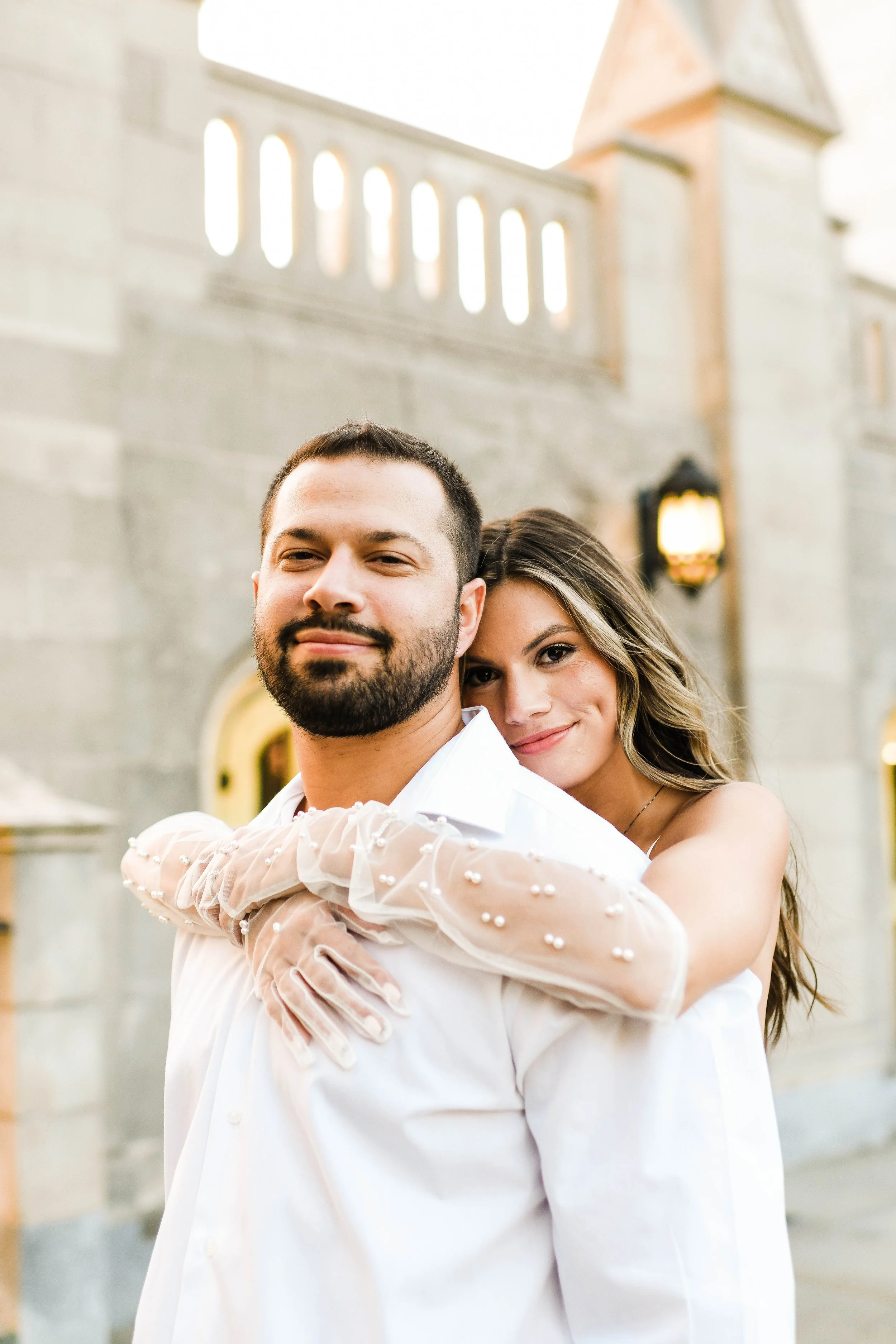 A couple in white clothing, with the woman hugging the man from behind, standing outside a stone building during sunset.