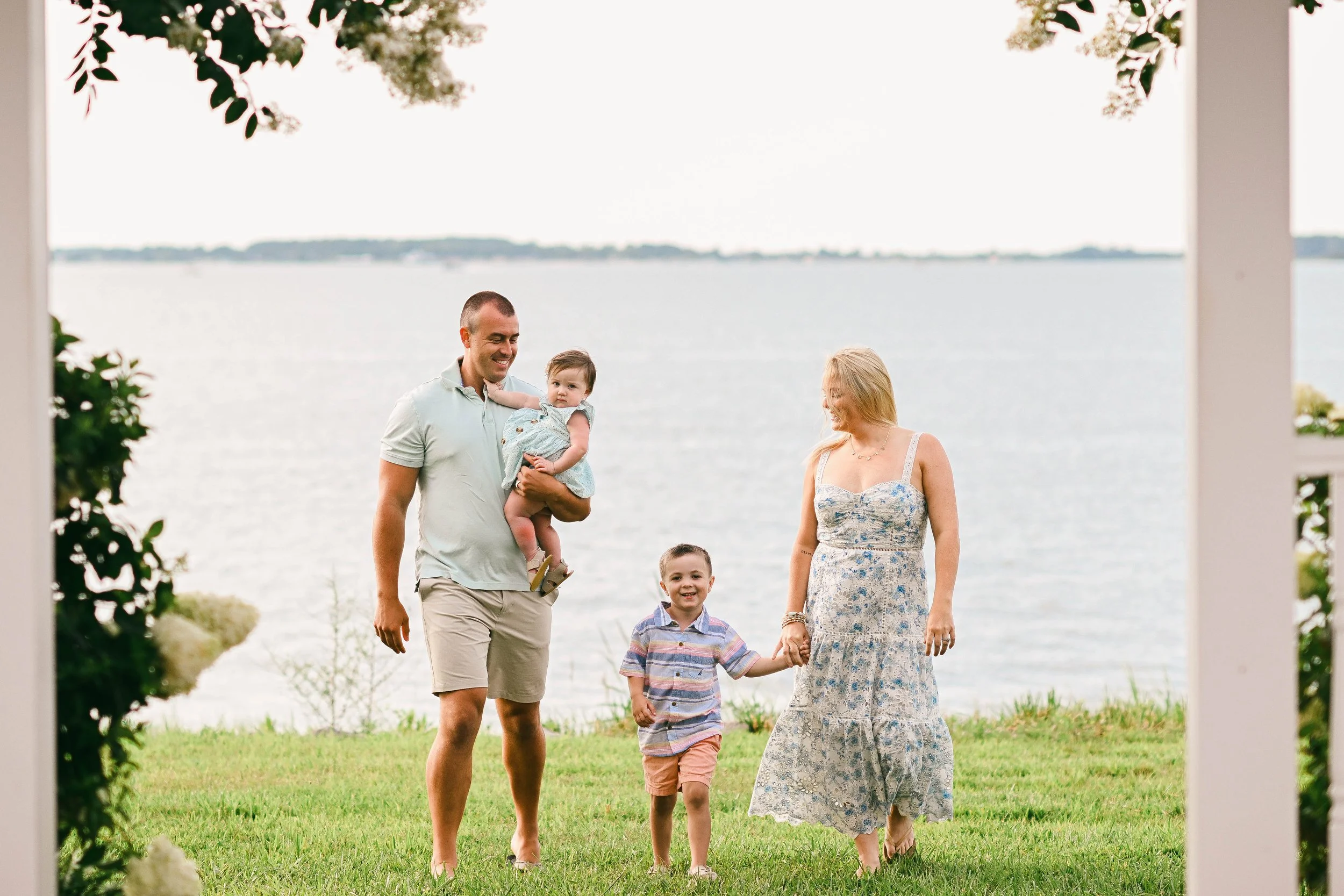 A family of four walking outside by a lake, holding hands, with trees around and a cloudy sky.