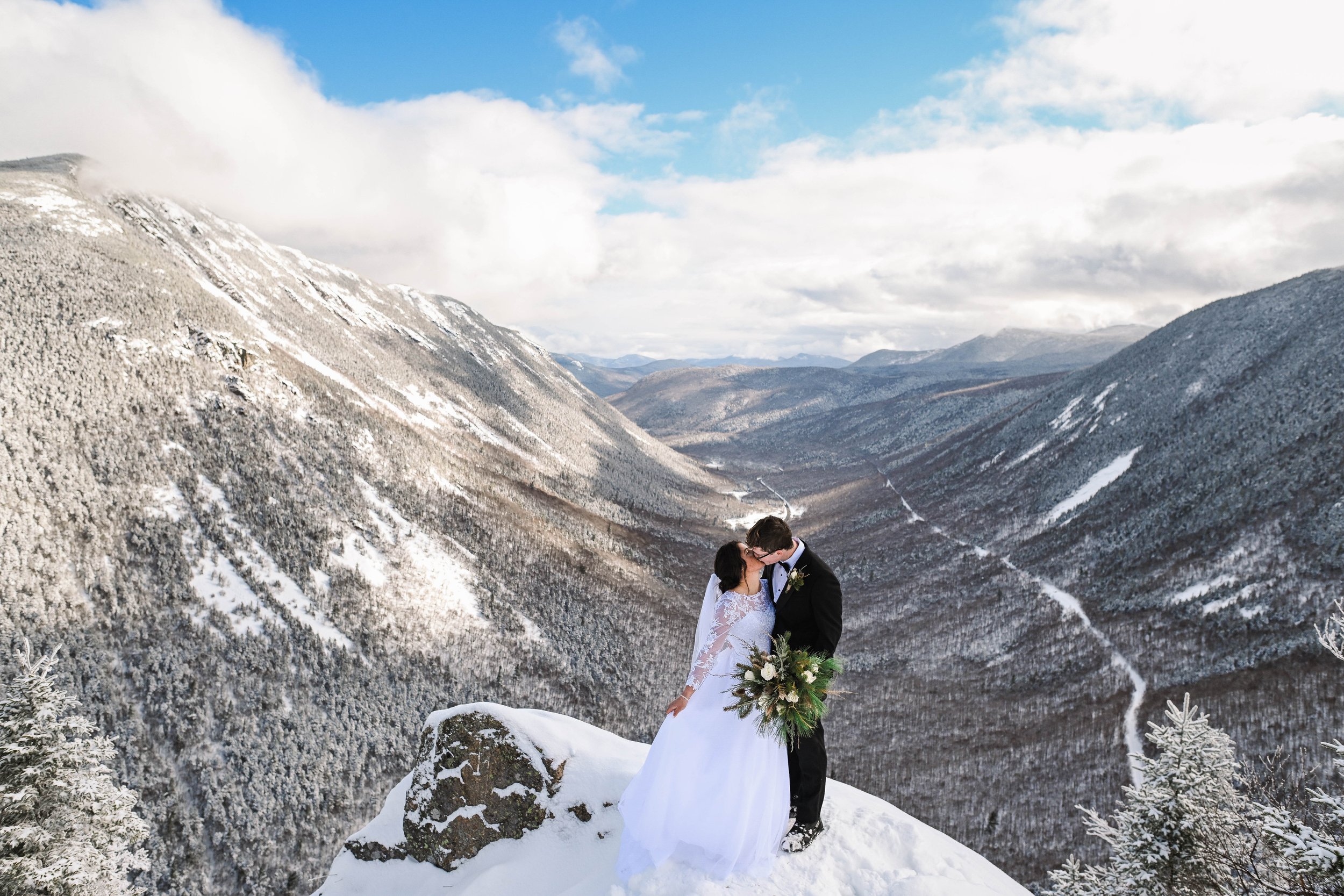 A newlywed couple in wedding attire sharing a kiss on a snowy mountain top with a scenic view of snow-covered mountains and a valley below.