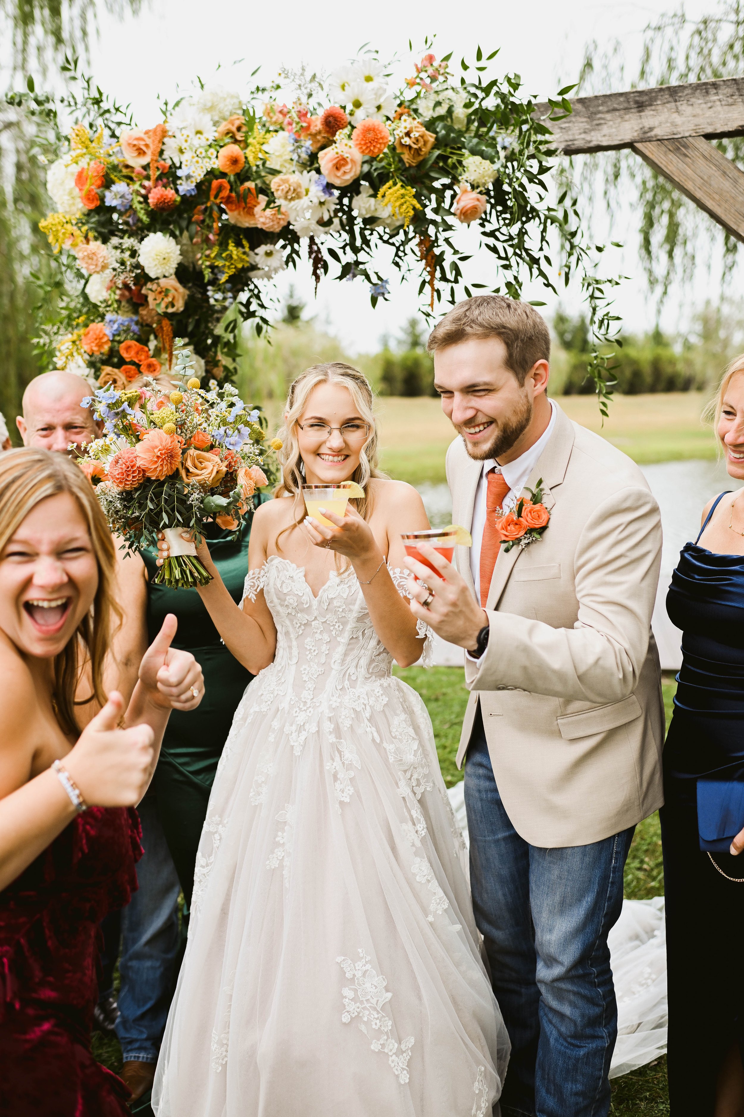 Group of people celebrating outdoors at wedding, with a bride in a white wedding dress holding a bouquet, and a groom in a beige suit, under a floral arch, enjoying drinks and smiling.