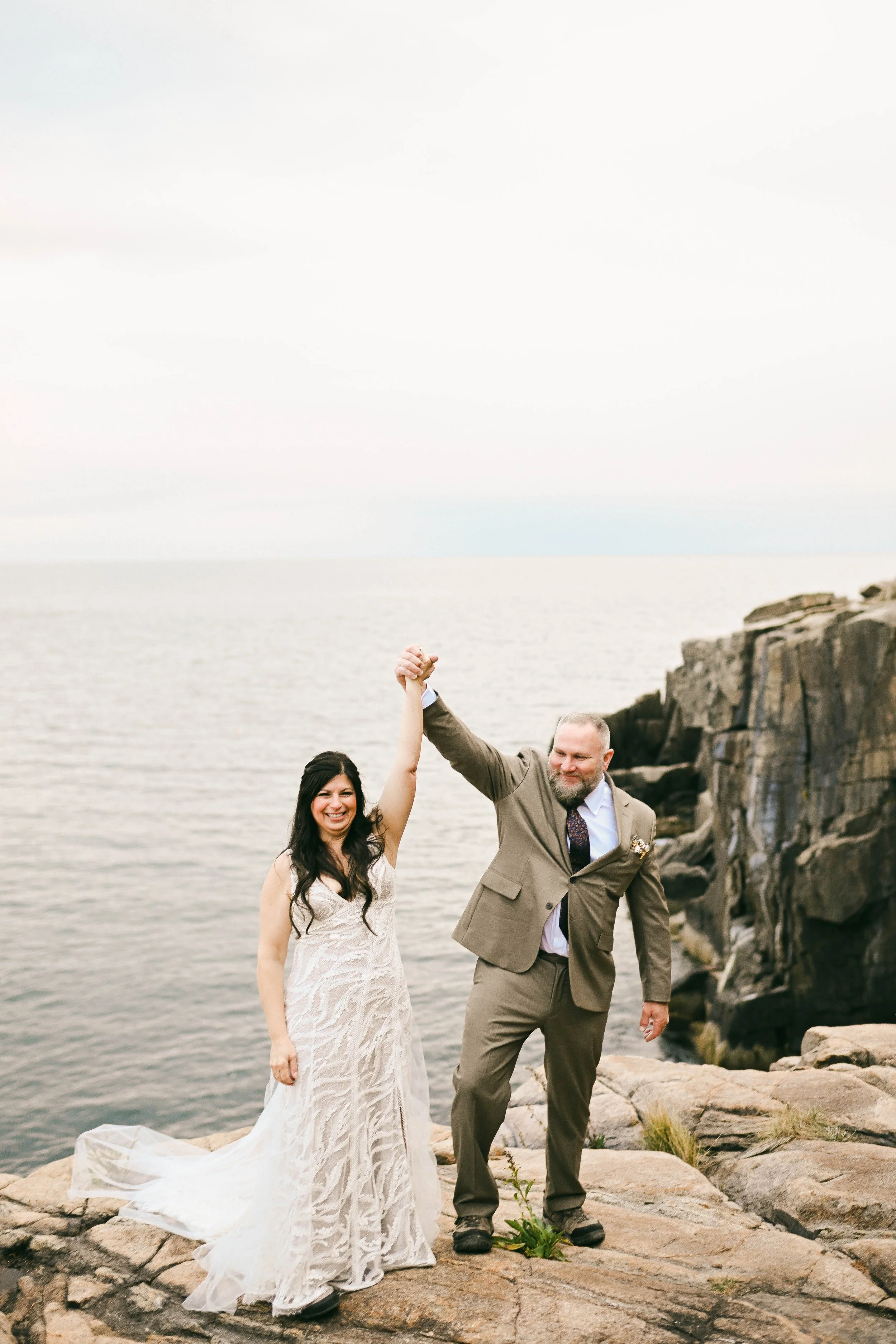 A bride and groom celebrating on a rocky shoreline by the water, holding hands in the air. The bride is wearing a white lace wedding dress and the groom is in a light brown suit.