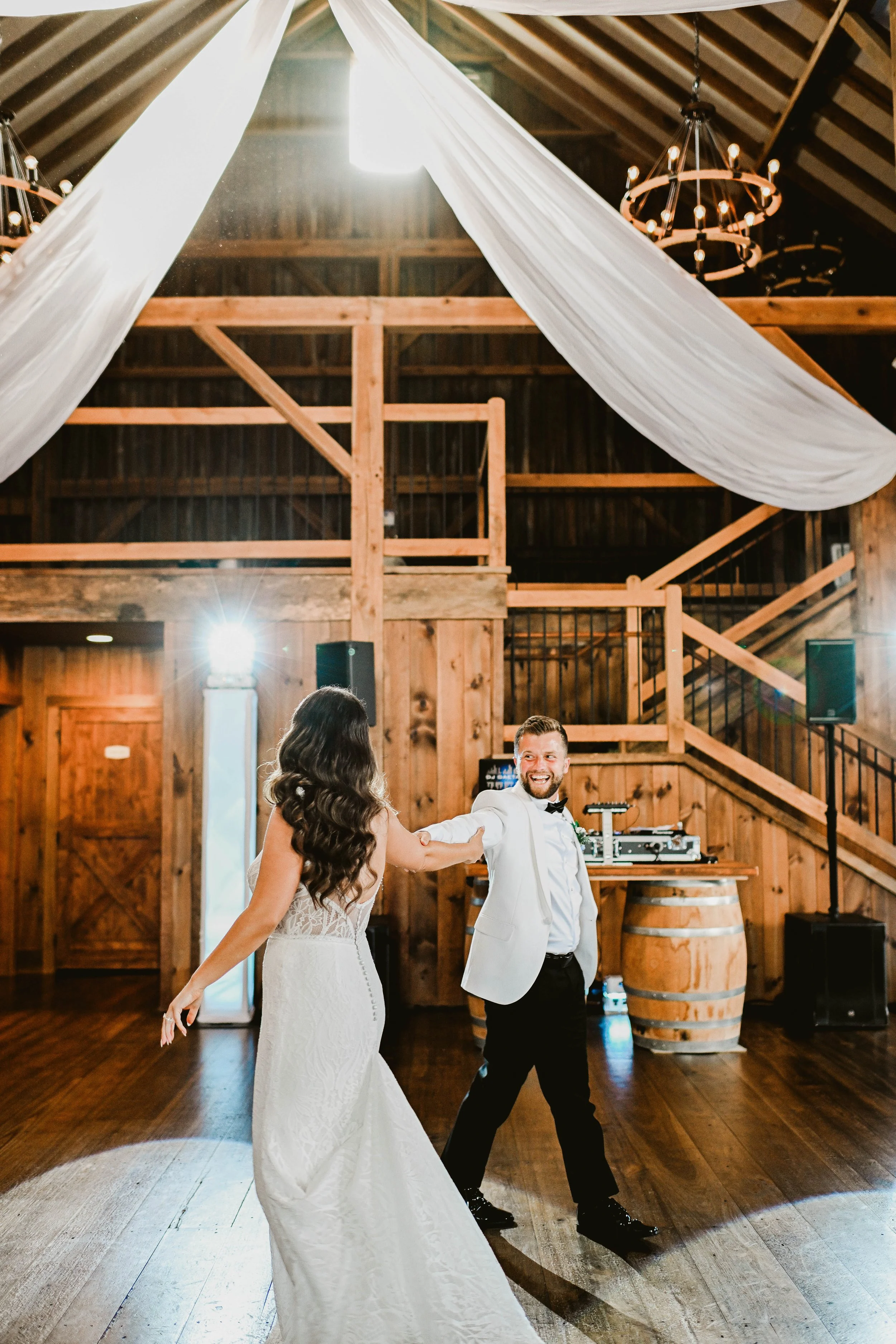 A bride and groom are dancing inside a rustic wooden barn decorated with large white drapes and chandeliers.