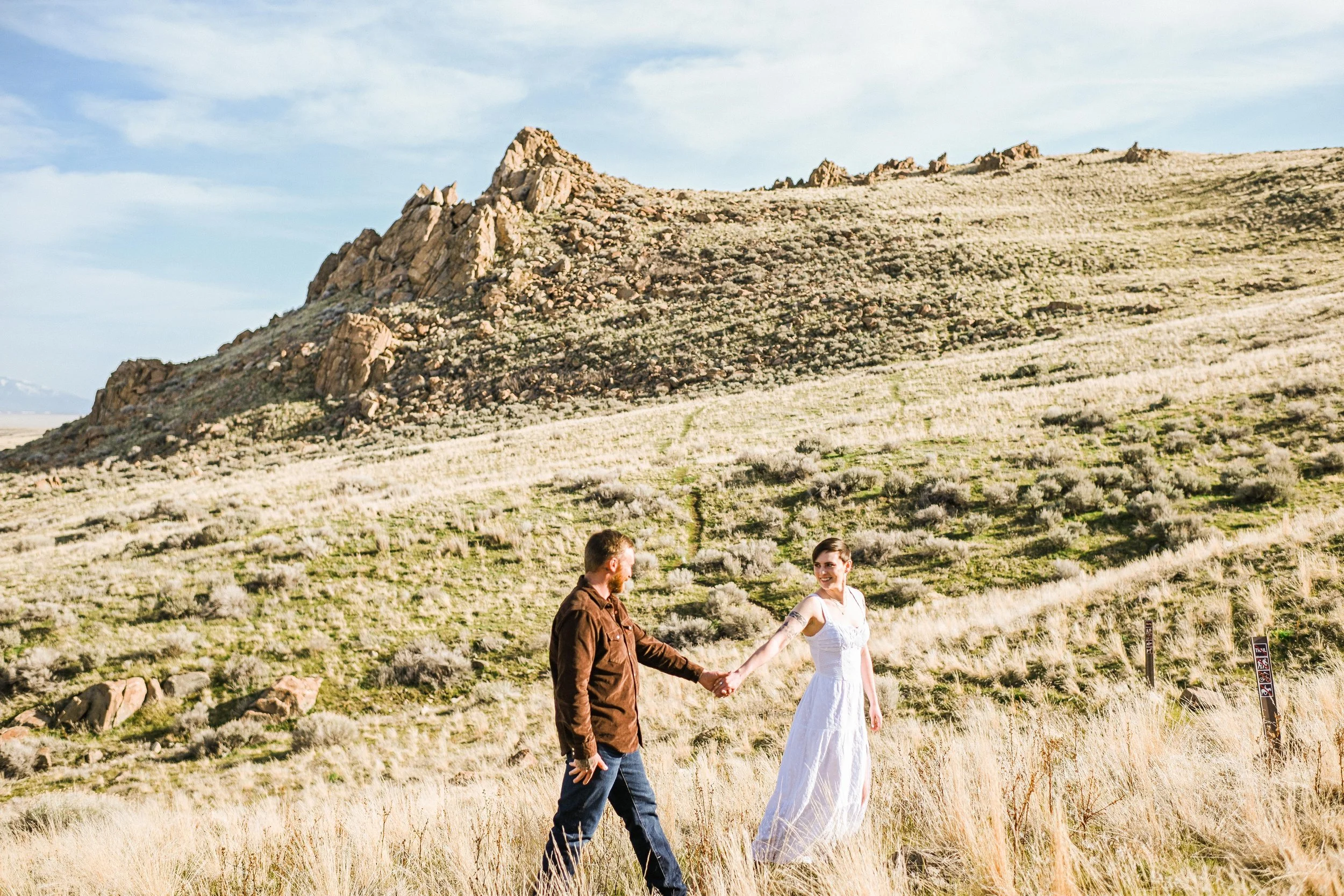 A couple holding hands in a grassy field with hills and rocks in the background, under a blue sky.