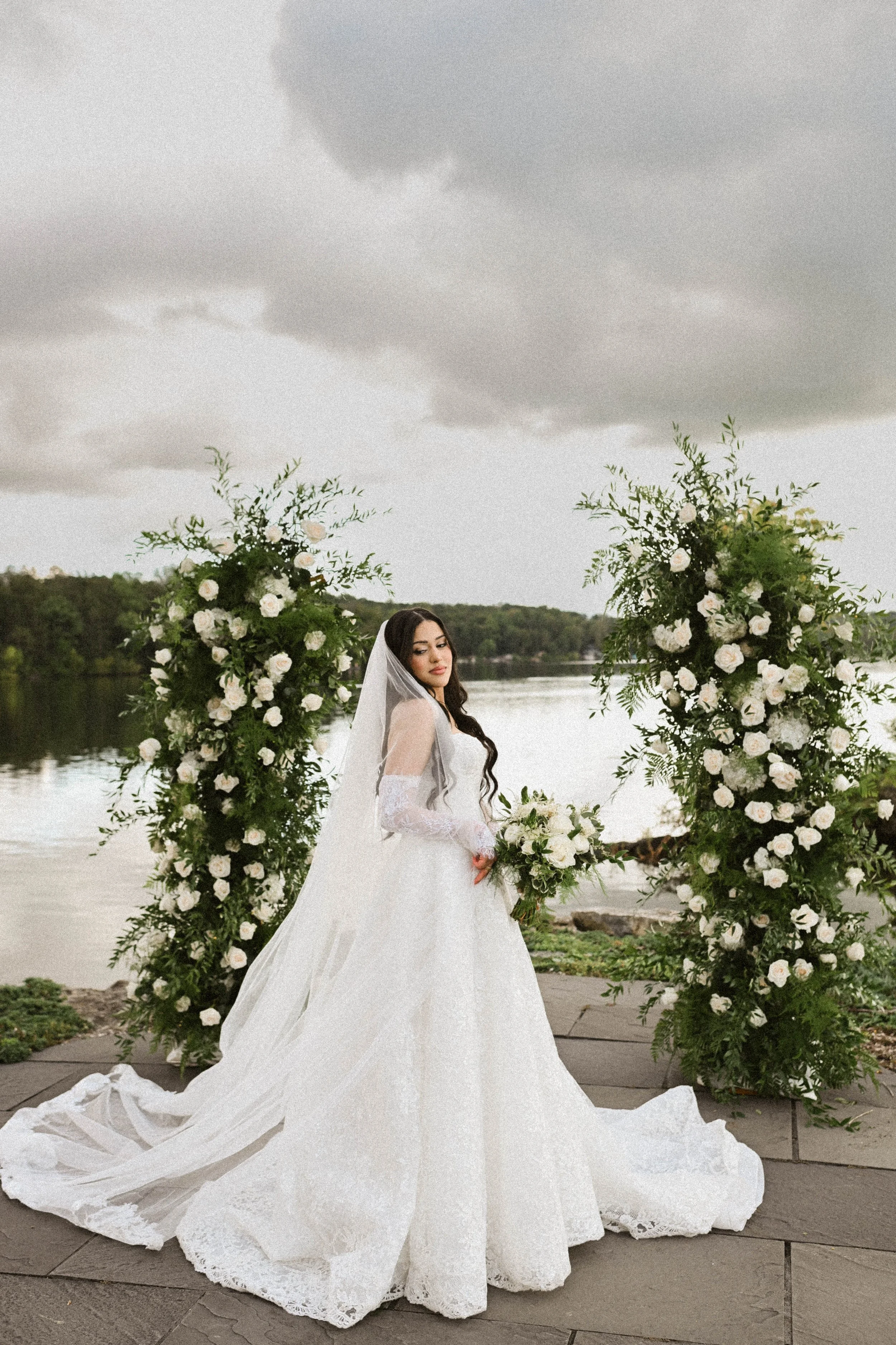 A bride in a white wedding gown with lace sleeves, holding a bouquet, standing outdoors near a river, with large floral arrangements on either side, under a cloudy sky.