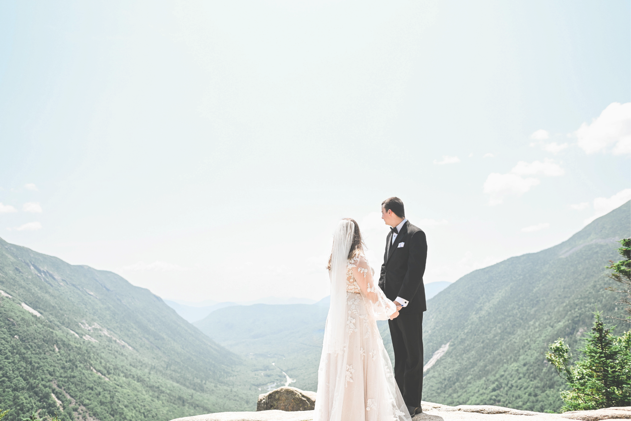 A bride and groom holding hands on a mountain overlook with green mountains and a bright sky in the background.