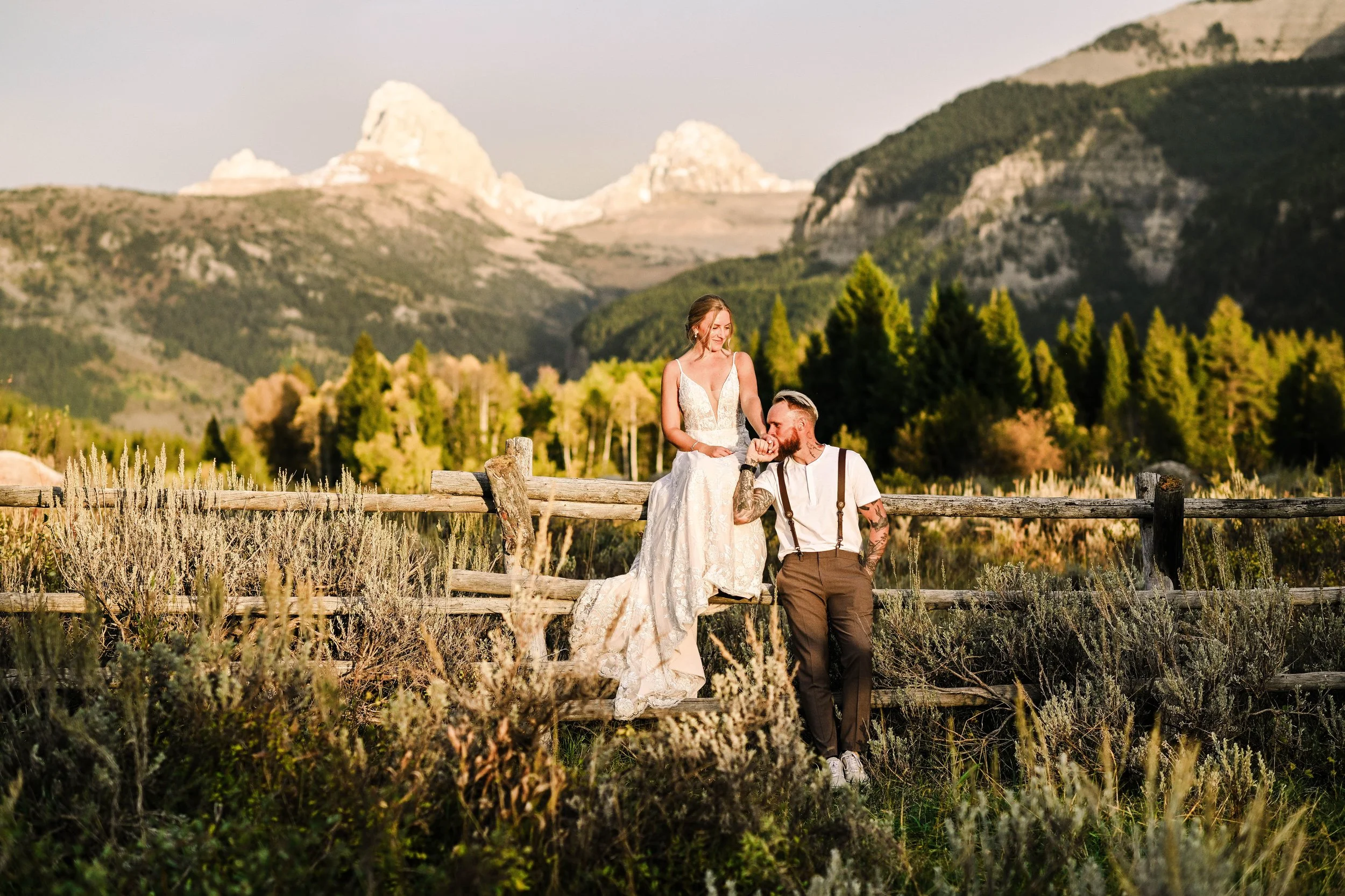 A couple with tattoos enjoying a romantic moment outdoors in a mountainous landscape, with the woman wearing a white dress standing on a wooden fence, and the man in a white shirt with suspenders and brown pants, kissing her hand against a backdrop of trees, hills, and mountains in the late afternoon.