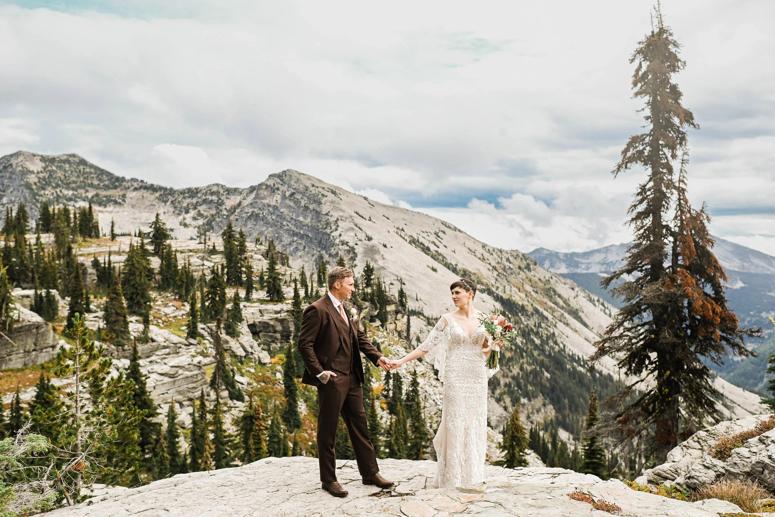 A bride and groom holding hands on a rocky ledge in a mountainous landscape with pine trees and cloudy sky.
