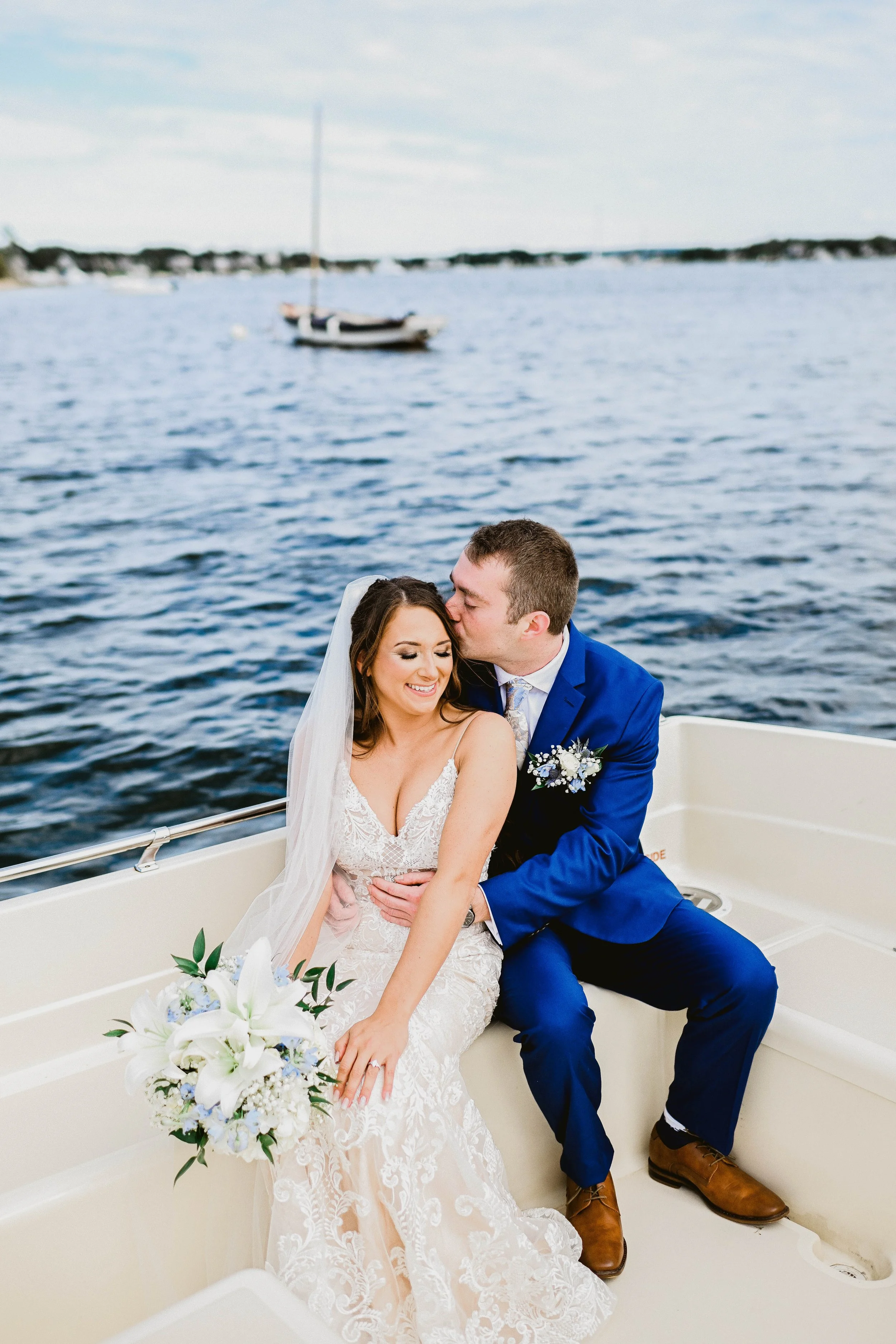 A newlywed couple sitting on a boat with a body of water and sailboats in the background