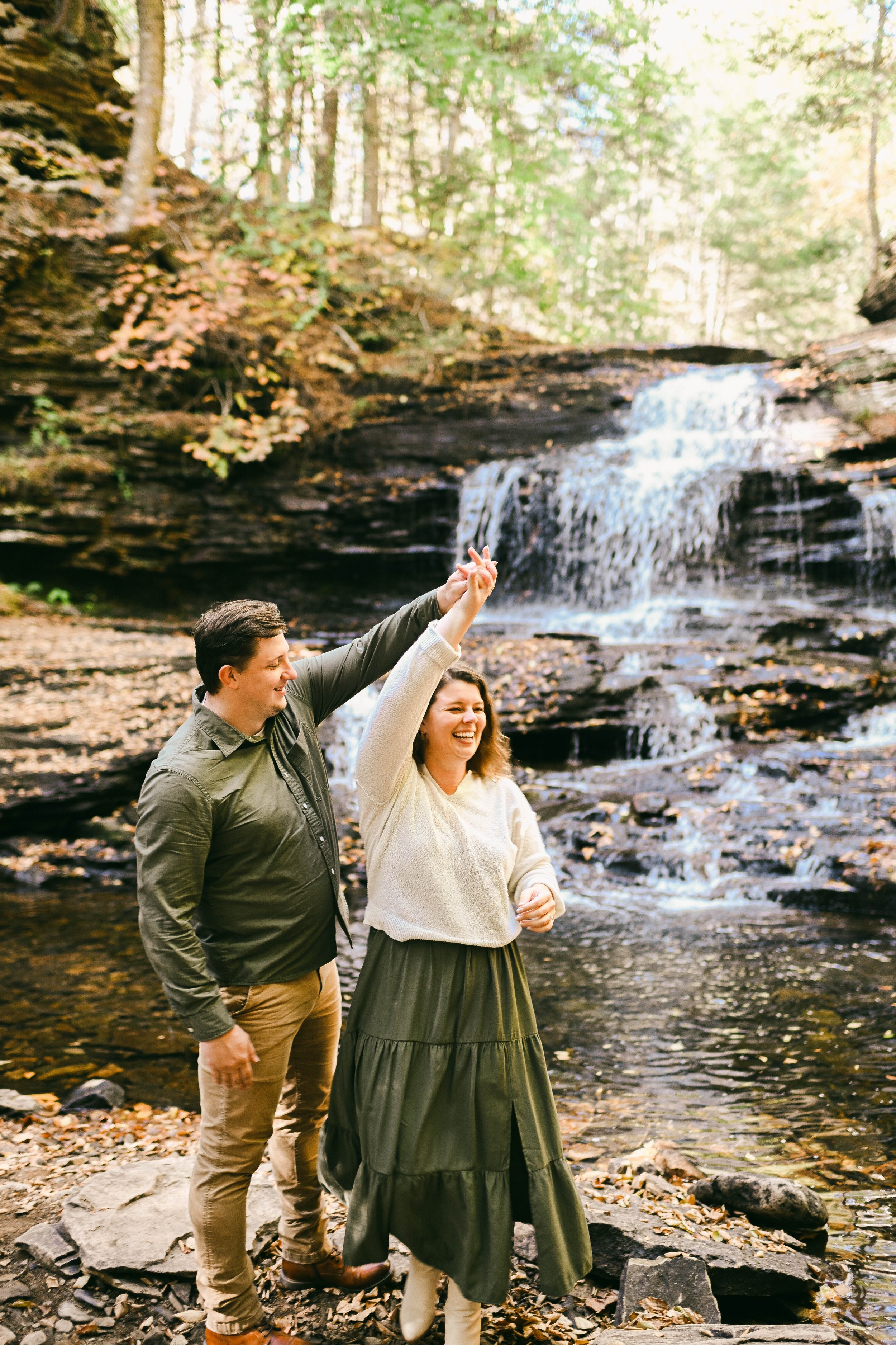 A smiling couple dancing and holding hands by a waterfall in a wooded area during autumn.