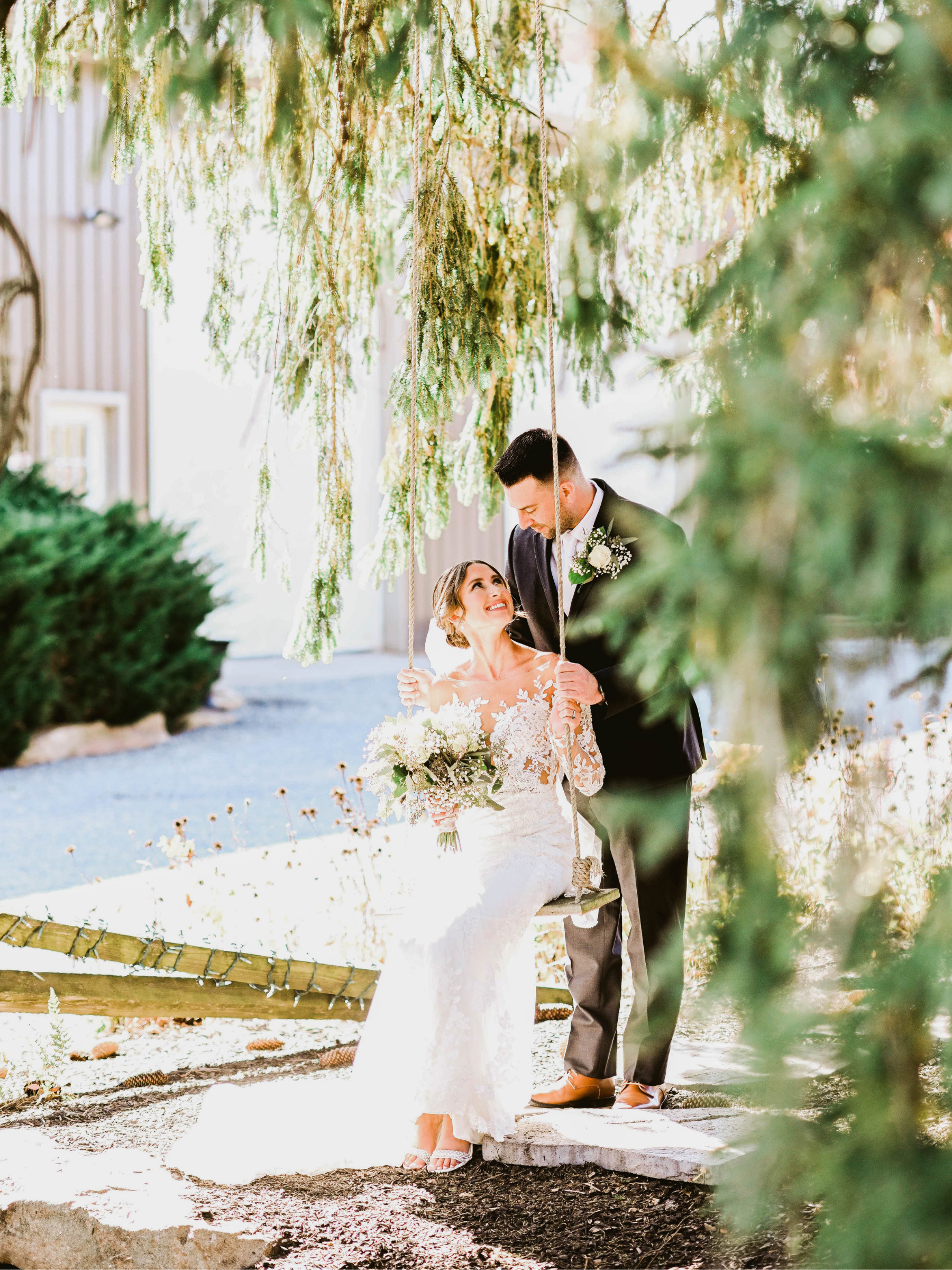 A bride and groom outdoors, the bride sitting on a swing holding a bouquet, looking up at the groom who is standing in front of her, both smiling. Greenery and hanging branches frame the scene with soft, natural sunlight.