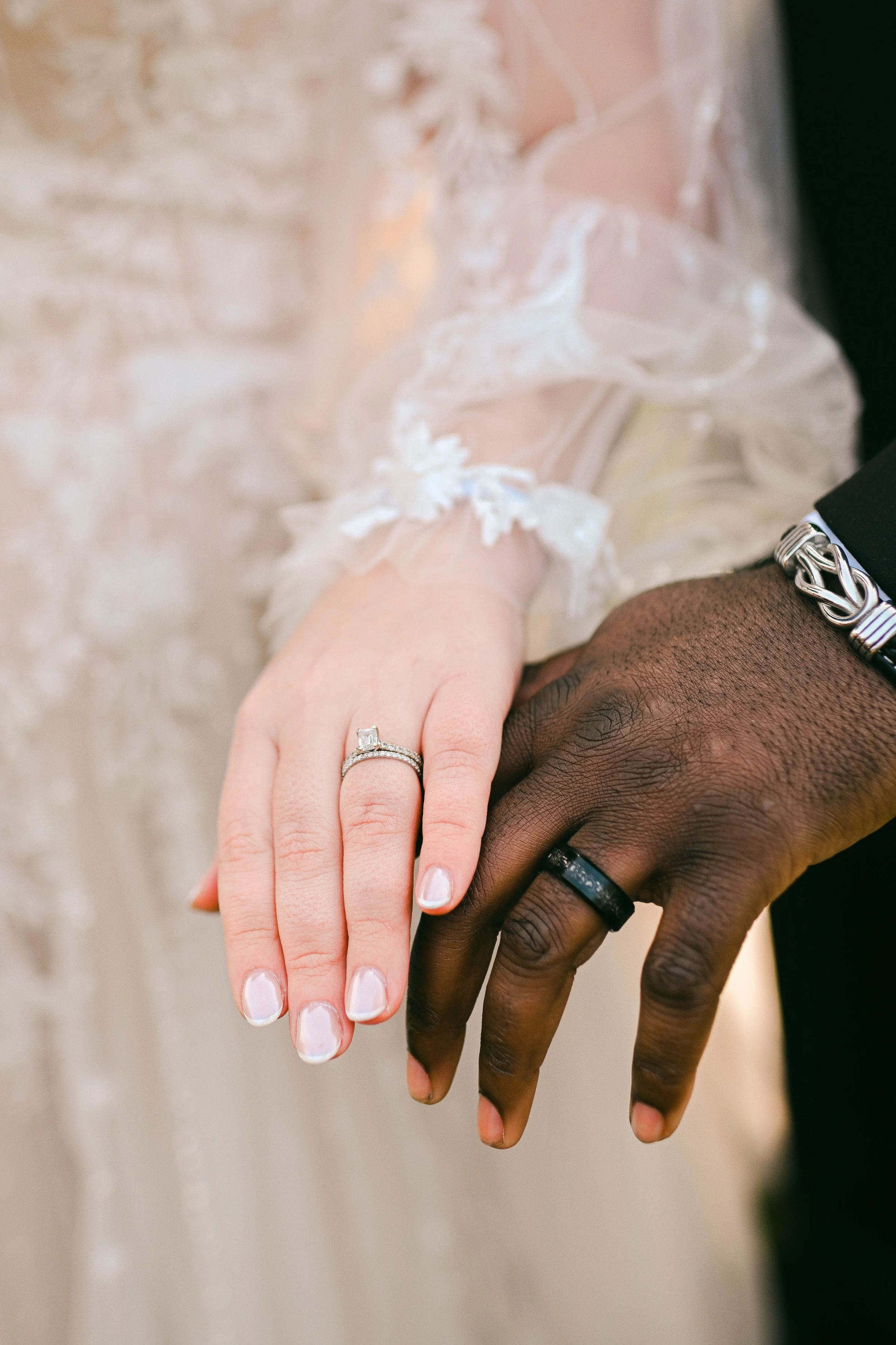 Close-up of a bride and groom holding hands. The bride wears a wedding ring and a sheer, lace-embellished sleeve, while the groom wears a black ring and a silver bracelet.