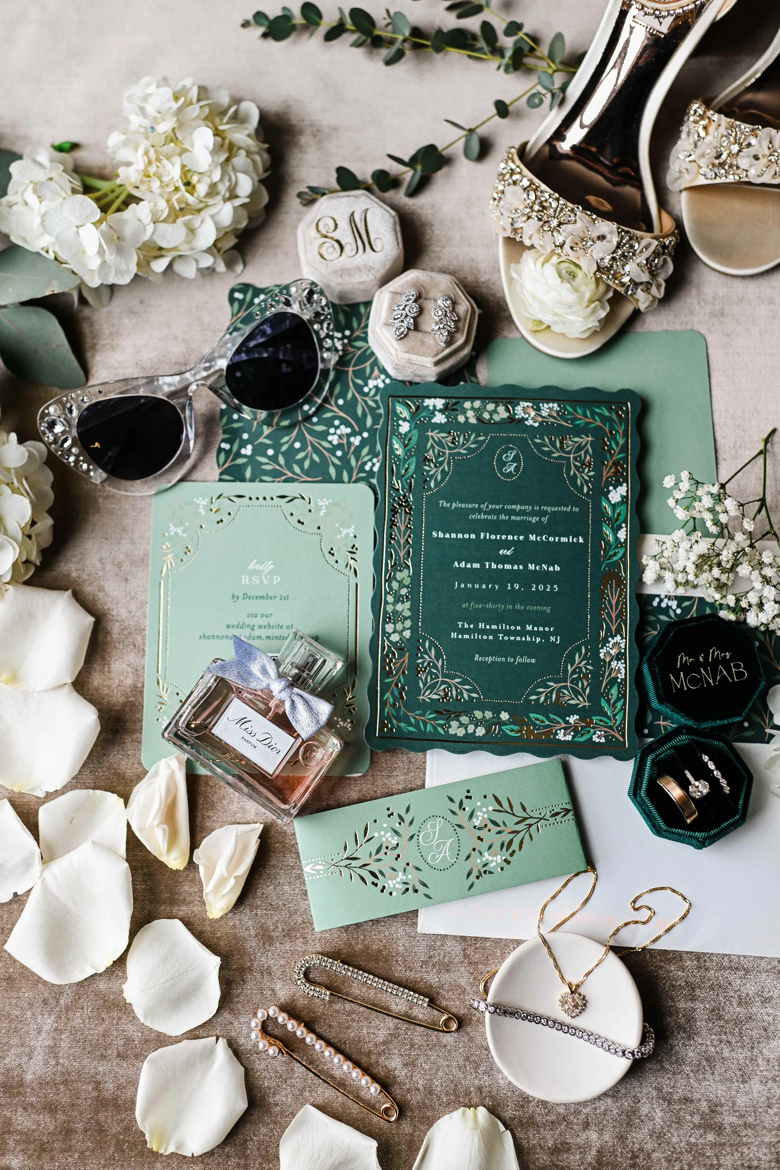 Flat lay of wedding items including floral accessories, jewelry, sunglasses, invitation cards, a perfume bottle, and shoes on a beige surface decorated with white rose petals and greenery.