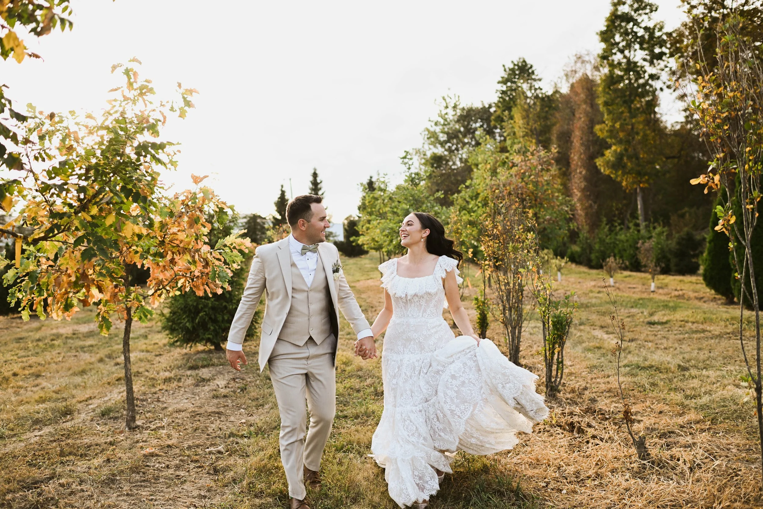 A newlywed couple holding hands and walking through a grassy outdoor area with trees in the background, both smiling at each other.