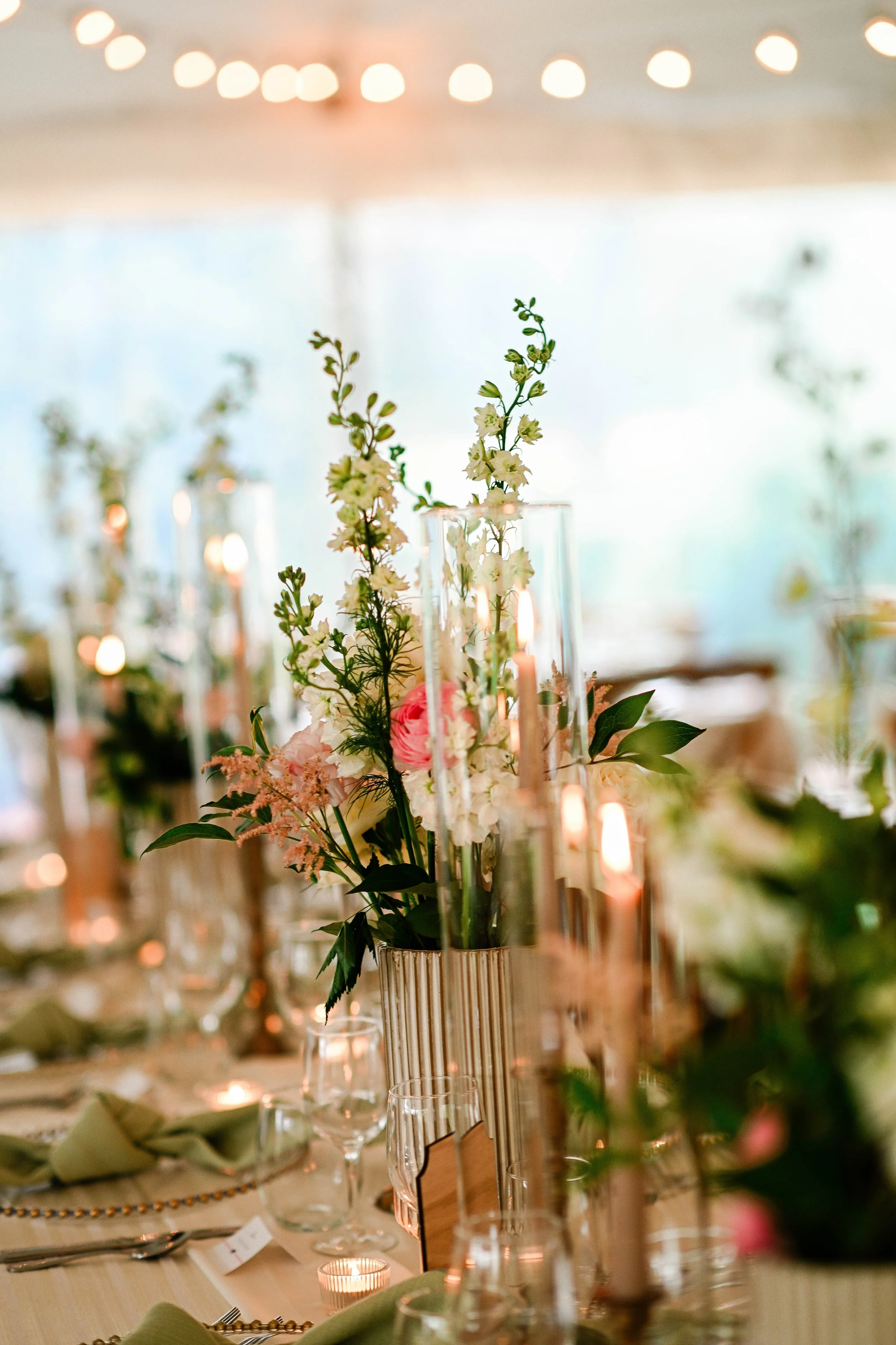 Elegant table centerpiece with white and pink flowers, surrounded by tall glass candle holders and lit candles, at a decorated event.