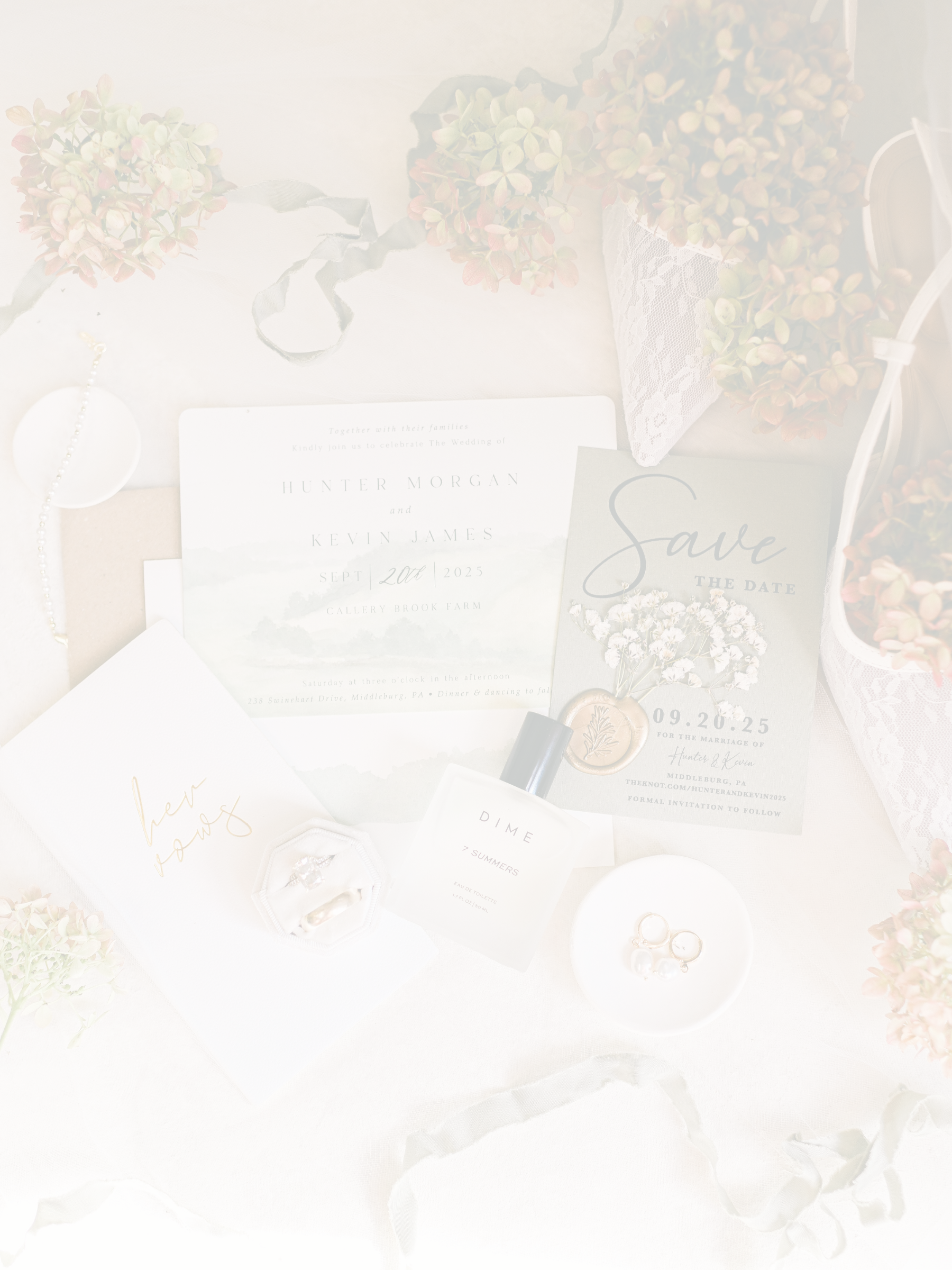 Wedding invitation, save-the-date card, wedding ring box with rings, white pearls, perfume bottle, jewelry, and floral arrangements on a soft fabric surface.
