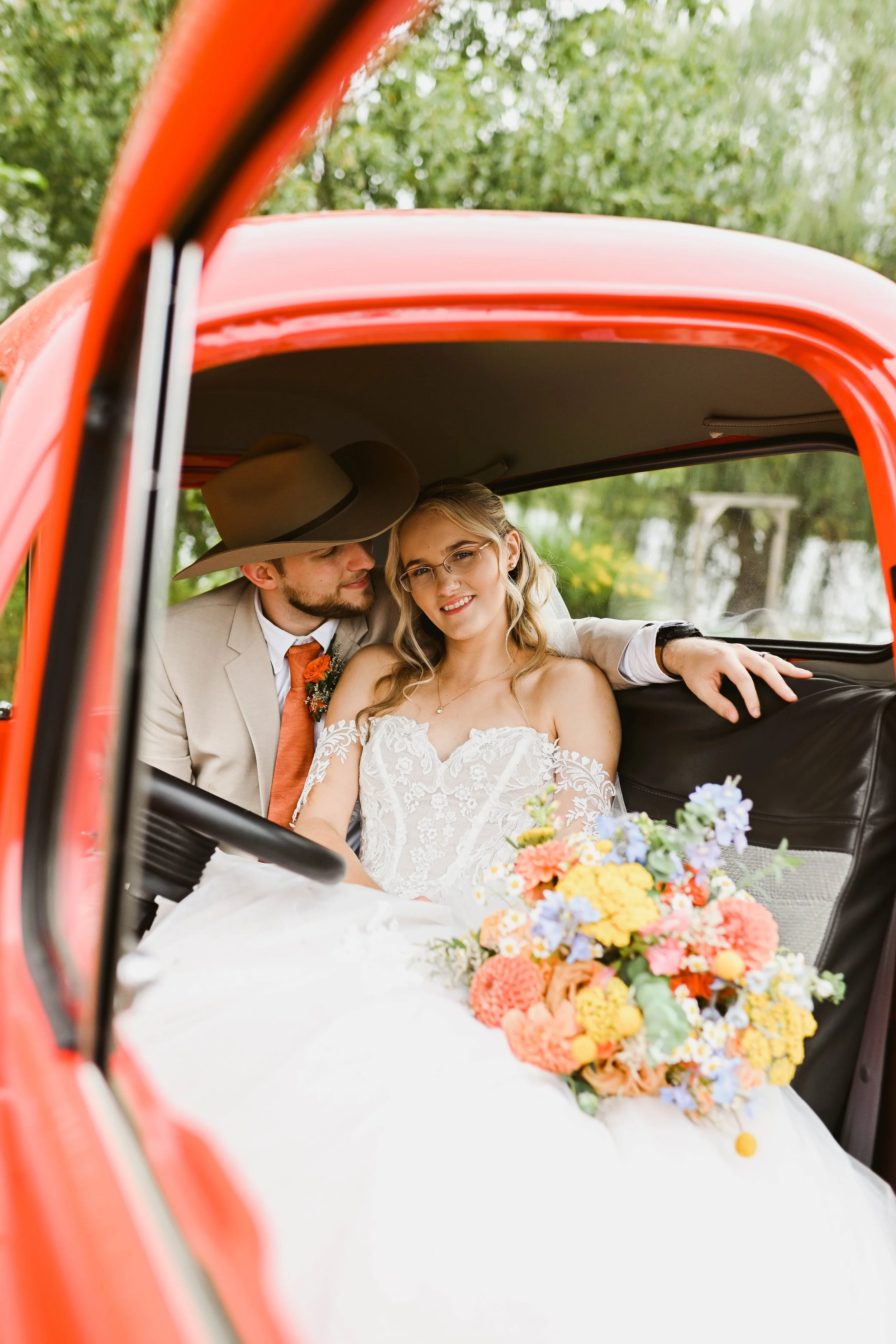A bride and groom sitting inside a vintage red car, with the groom whispering to the bride, who is smiling. The bride is in a white lace wedding dress with a floral bouquet on her lap.