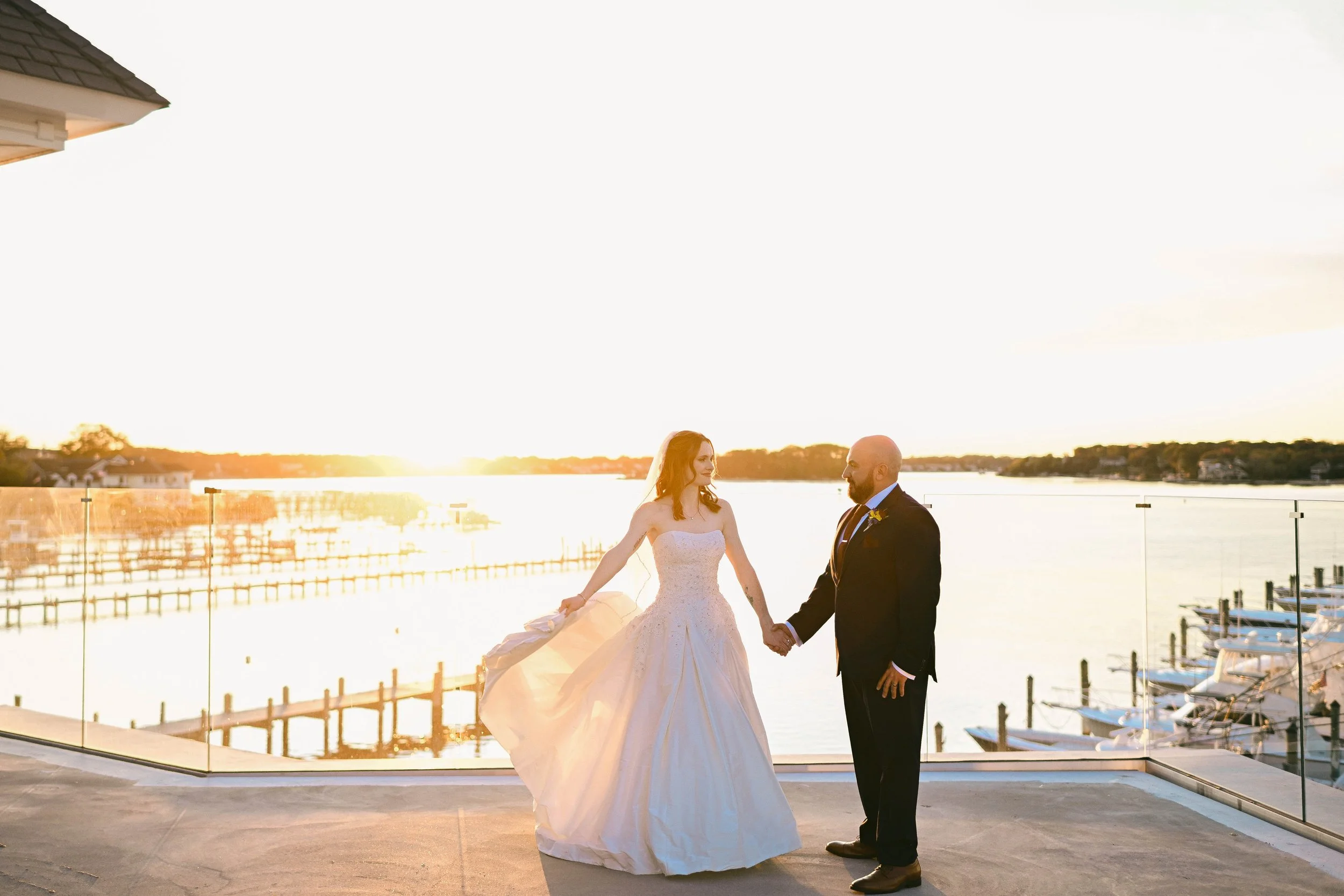 A bride and groom holding hands on a balcony overlooking a marina at sunset.