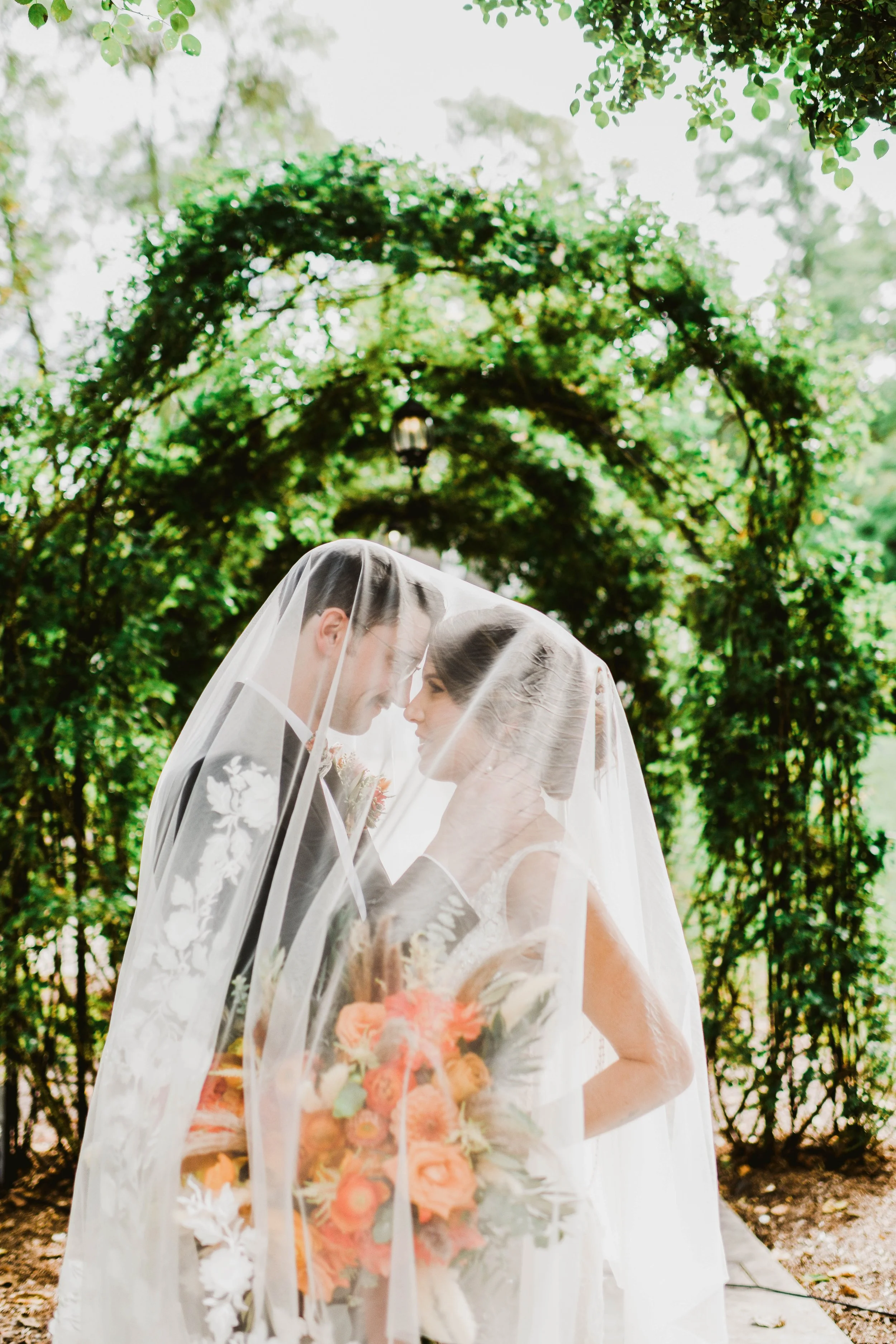 A bride and groom stand close together under a floral arch, sharing a moment covered by a bridal veil, with greenery in the background.