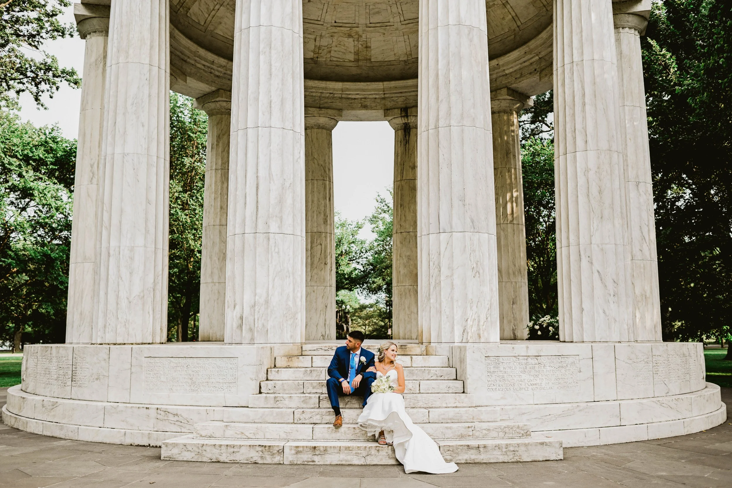 A bride and groom sitting on steps of a white marble monument in a park, with trees in the background.