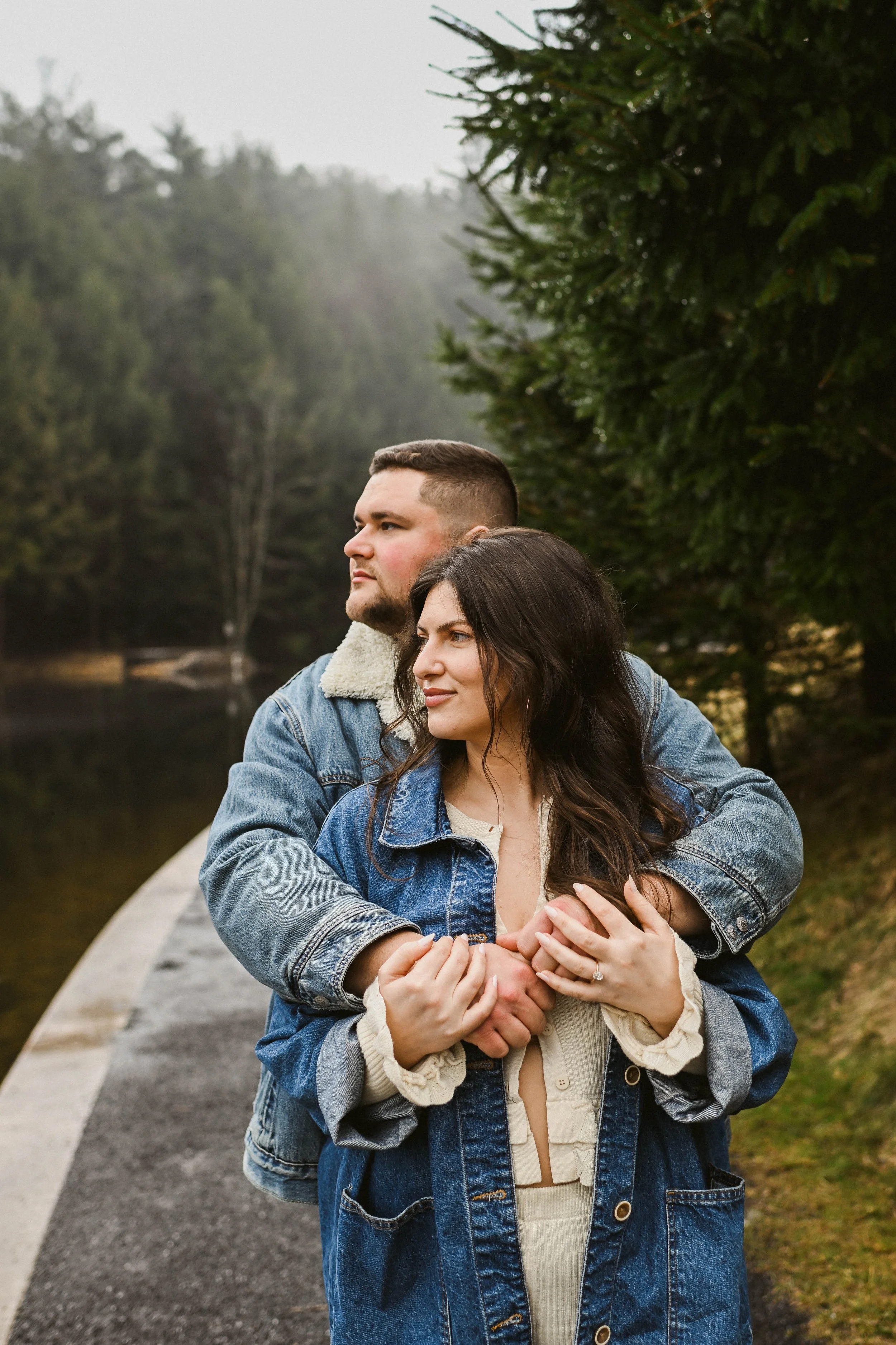 A couple standing outdoors on a cloudy day, with the man hugging the woman from behind near a lake and trees.