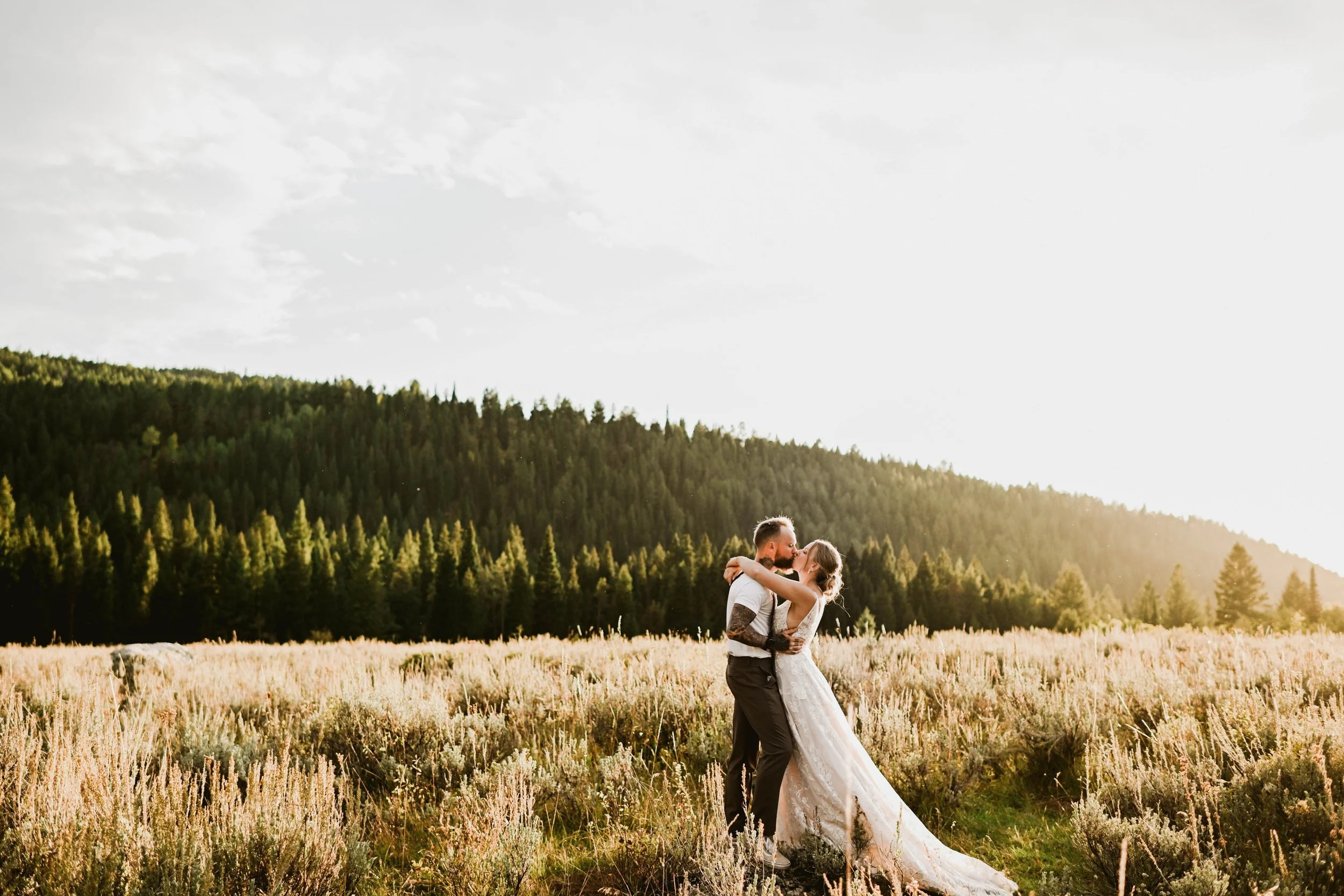 A couple in wedding attire embracing and kissing in a field with a forested hill in the background, illuminated by sunset light.