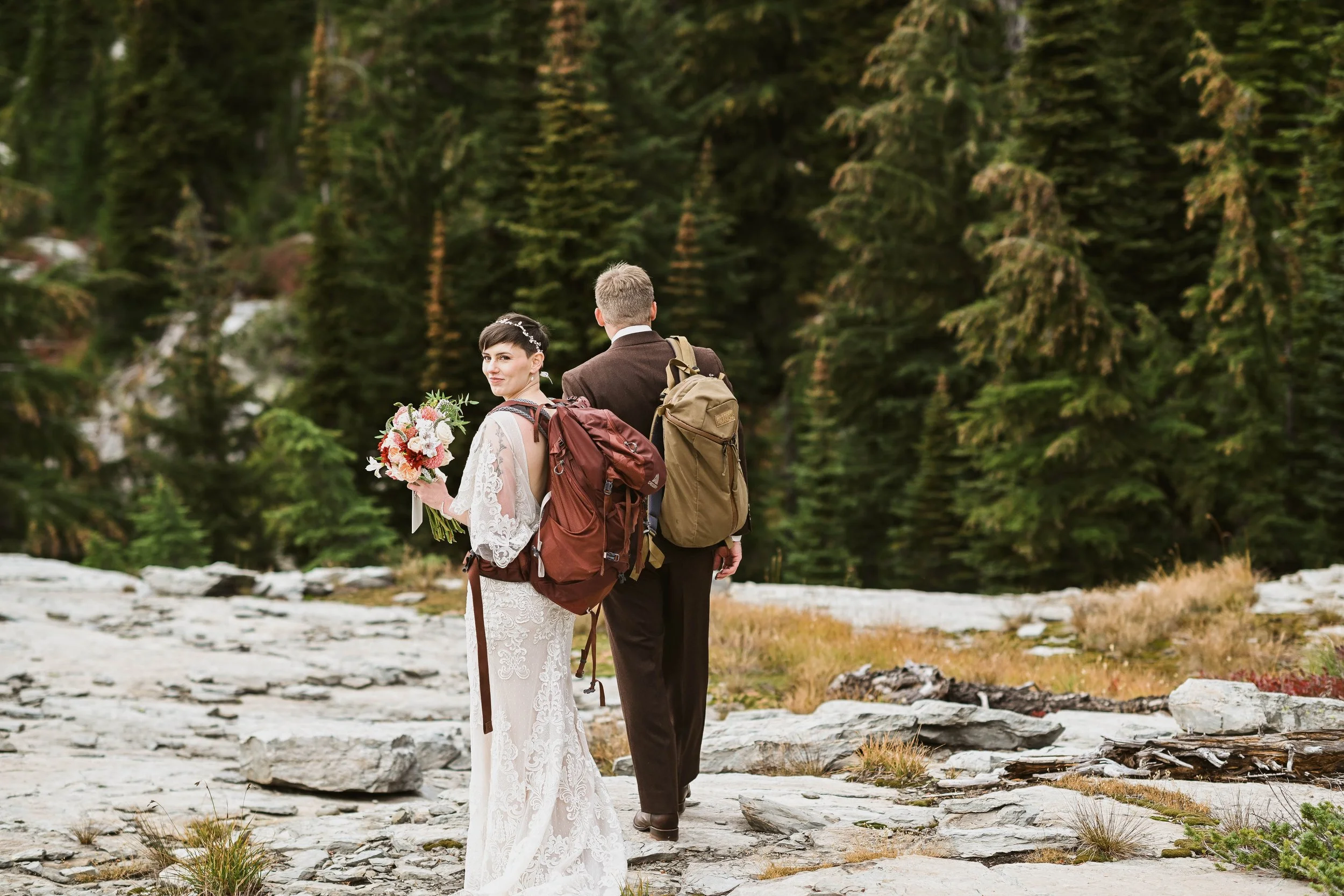 Bride and groom with backpacks standing on rocky terrain in a forested area.