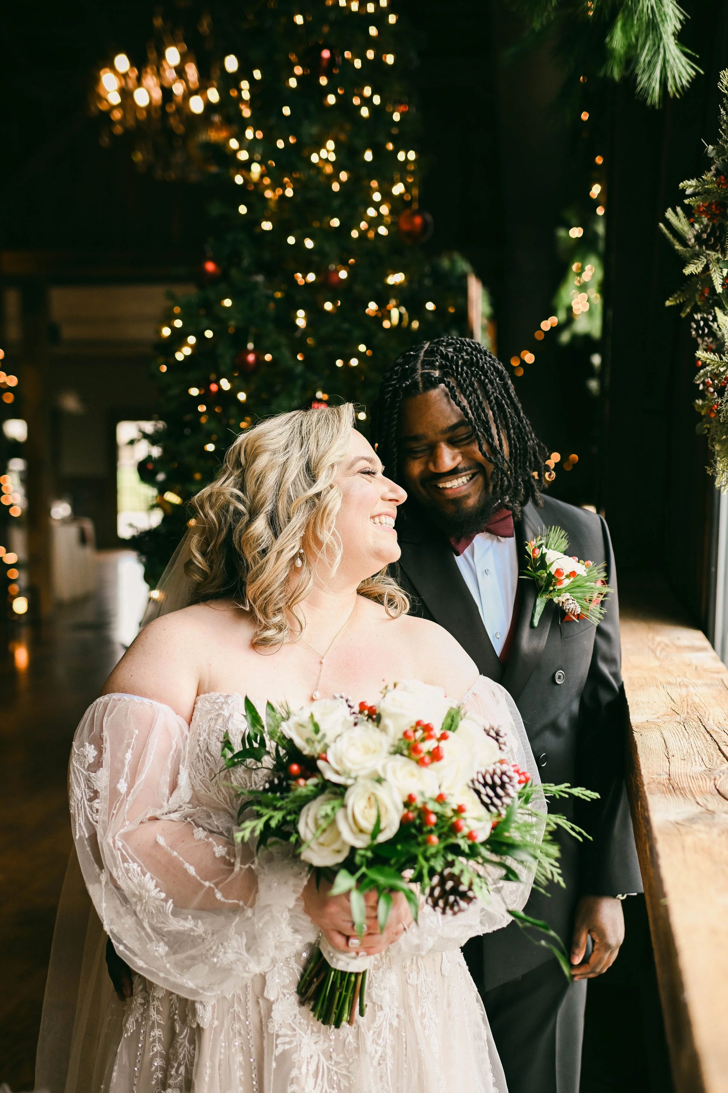 A bride and groom smiling joyfully, standing beside a window with holiday decorations, including a Christmas tree with lights and ornaments, in a warmly decorated indoor setting.