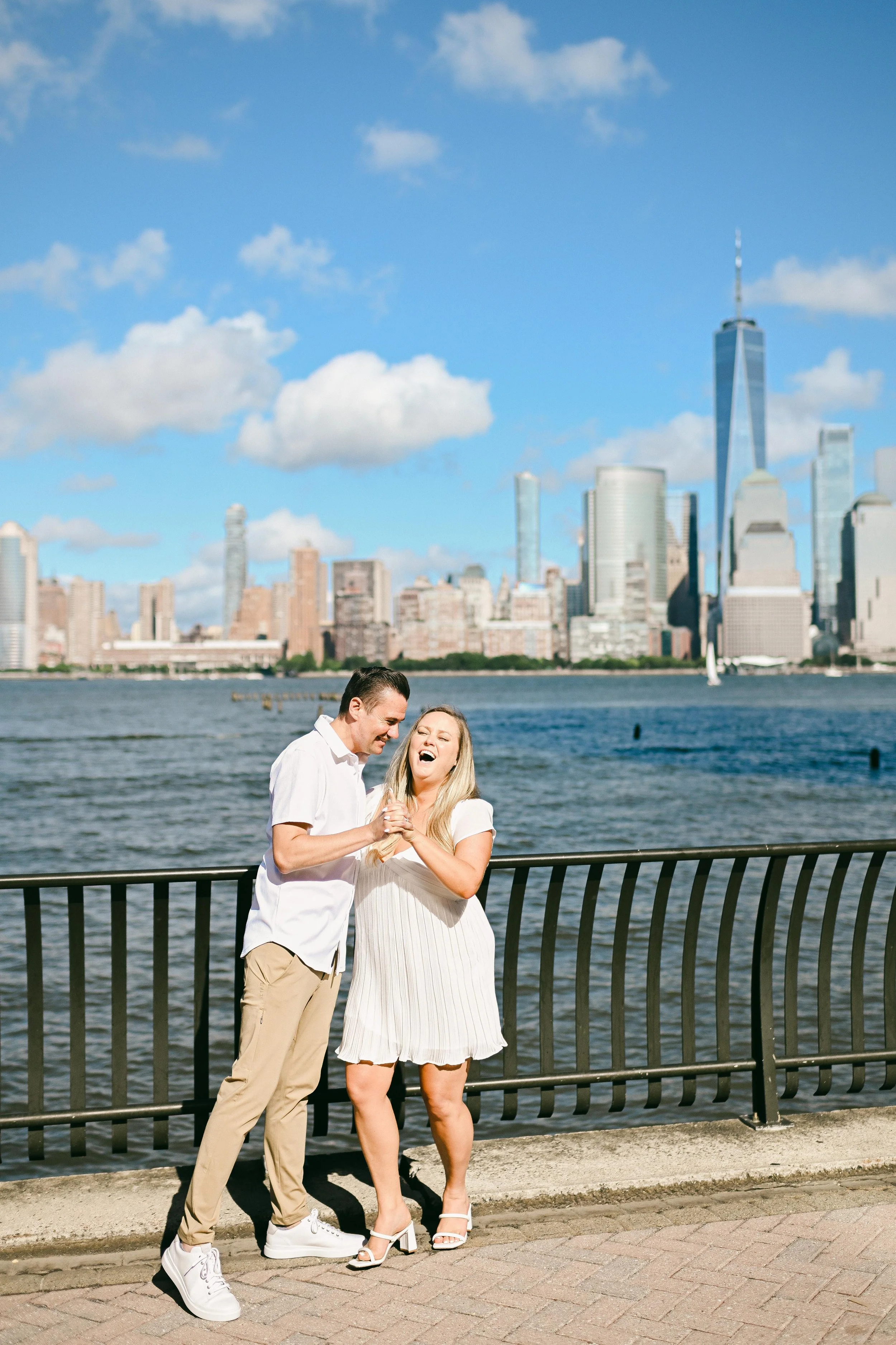 A couple laughing and holding hands, standing by a waterfront with a city skyline in the background, including a tall building with a spire, under a blue sky with fluffy clouds.