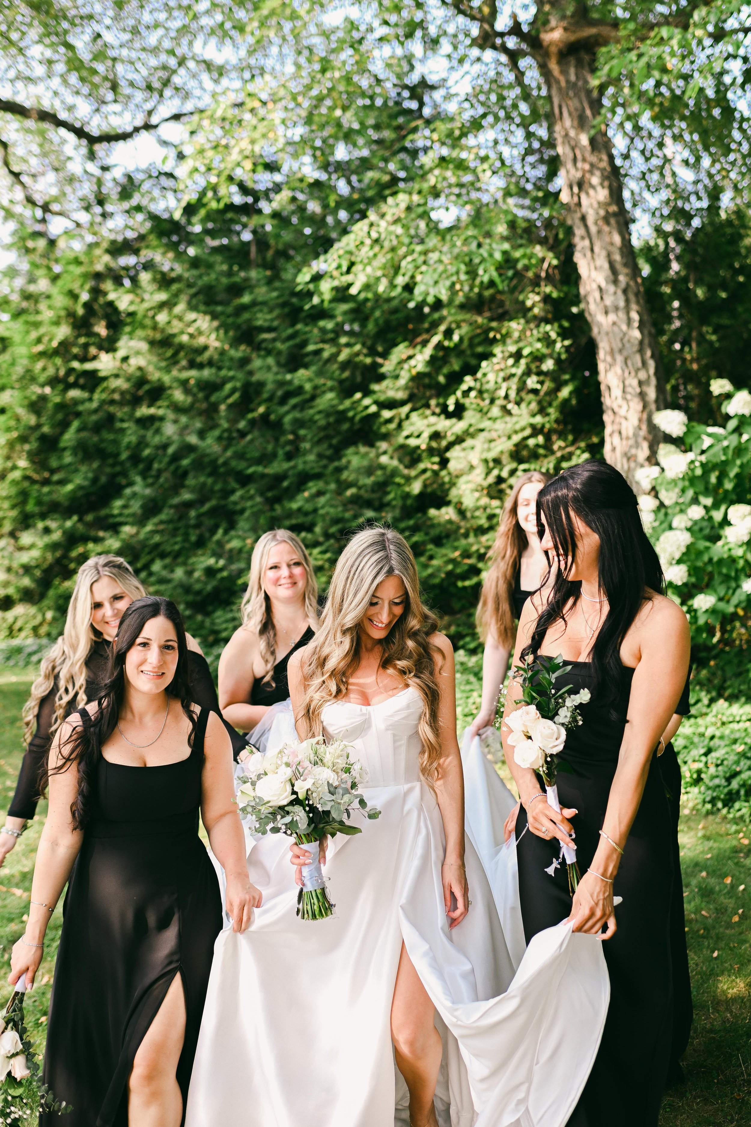Group of women, including the bride in a white wedding dress, walking outdoors among trees and green foliage, holding bouquets and smiling.