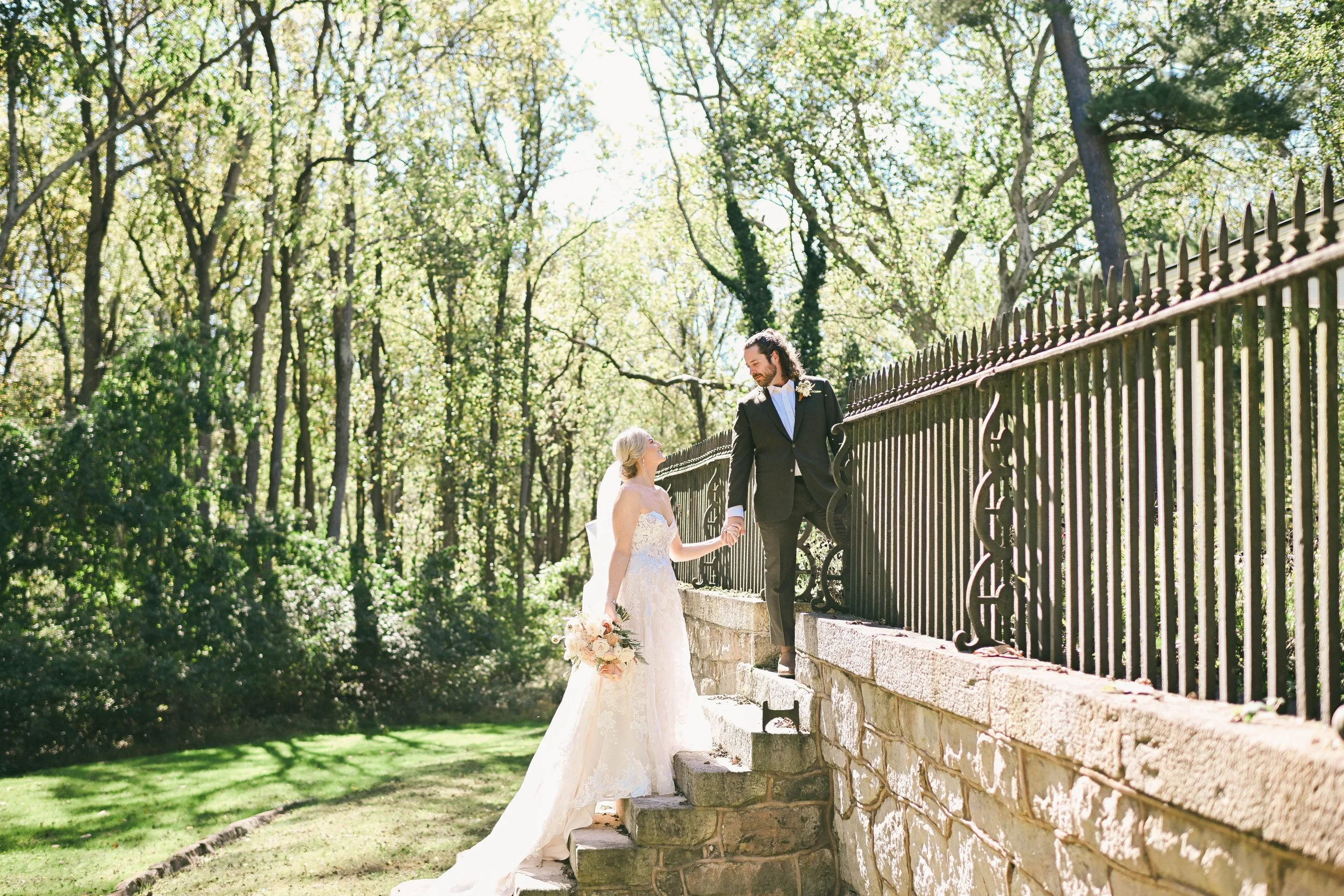 A bride and groom holding hands on outdoor stone stairs beside a wrought iron fence, with trees and sunlight in the background.